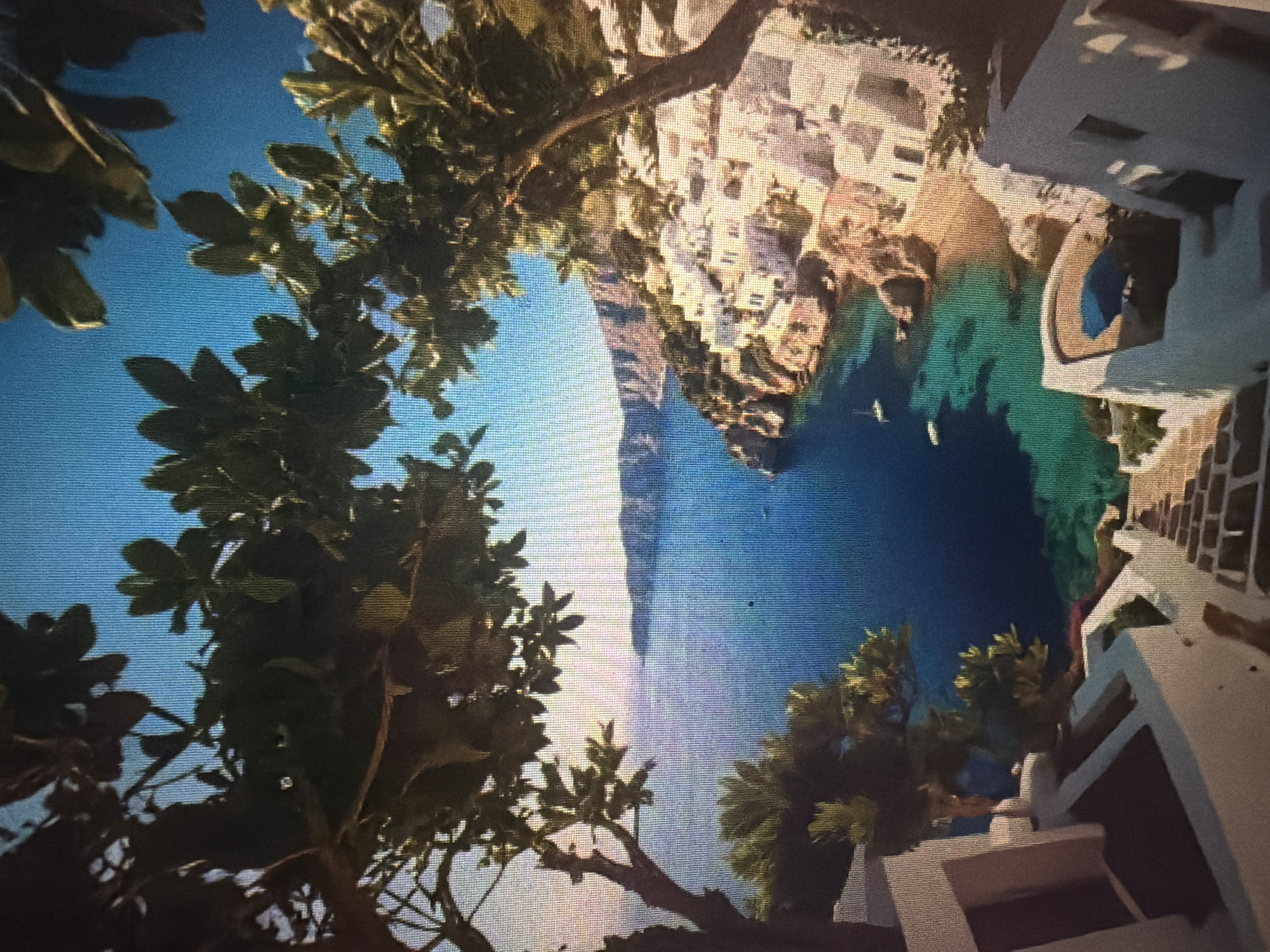 This is a vertical, medium-shot photograph taken from a high vantage point, looking down onto a coastal village and the sea. In the foreground, white, cube-shaped buildings with flat roofs, characteristic of Mediterranean architecture, cascade down a hillside. A stone staircase leads down between these buildings. Lush green foliage, with dark green leaves and branches, frames the top and sides of the image, obscuring parts of the sky and the village.
The village itself is a cluster of white buildings built into a rocky incline that meets the sea. The buildings have simple, clean lines and some appear to have small windows and doors. Below the village, a small, sandy cove is visible, with clear, turquoise water lapping at the shore. The sea stretches out to the horizon, where it meets a hazy, mountainous coastline under a bright, clear blue sky. Two small white sailboats are visible in the water. The overall scene evokes a sense of tranquility and picturesque beauty.
The image does not depict the city of Baan Mae Nam, Thailand. The architectural style and landscape are characteristic of a Greek island, such as Santorini or Mykonos, or other similar Mediterranean coastal regions.