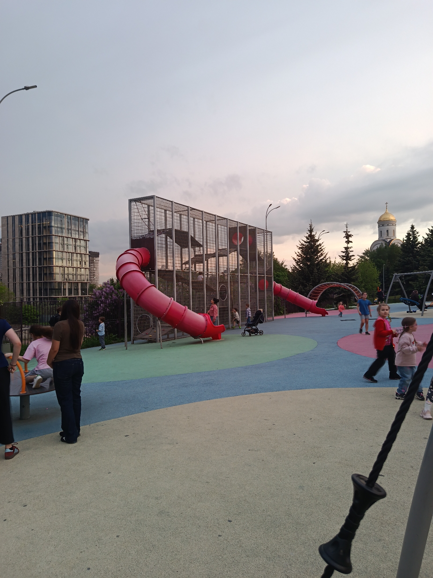 The image depicts a children's playground in Moscow, Russia. In the background stands a building with a grid-like facade and the golden dome of a Russian Orthodox church. In the center of the playground is a tall play structure with a red tube slide. A few children are near the play structure and others are scattered across the colorful playground surface. Several adults are standing nearby. The sky is overcast.
