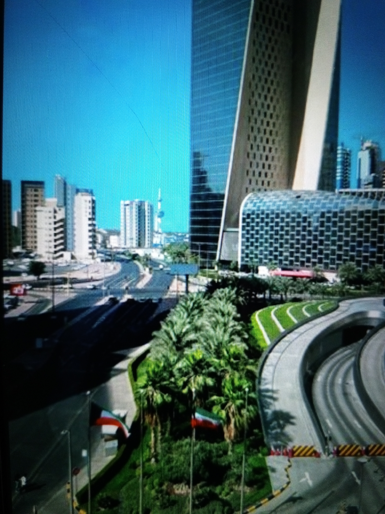 The image shows a cityscape with modern architecture under a clear blue sky. A prominent, tall, angular building with a textured facade dominates the right side of the frame, appearing to be a skyscraper. To its left, another, lighter colored skyscraper with a smooth surface is visible, partially obscured. Further to the left, a cluster of various buildings, including what appears to be another skyscraper with a patterned glass facade, are visible. In the foreground, a multi-lane highway curves through the scene, with sections of overhead walkways or bridges. Greenery, including palm trees, is interspersed throughout the urban landscape, particularly along the highway's curves. Two flags, one with red, white, and green horizontal stripes, and another with red, white, black, and green elements, are visible on poles in the lower left and lower right of the image respectively. The overall impression is of a well-developed, modern city. The location in the city of Golbembal, Nigeria, is not discernible from the visual elements in the image.
