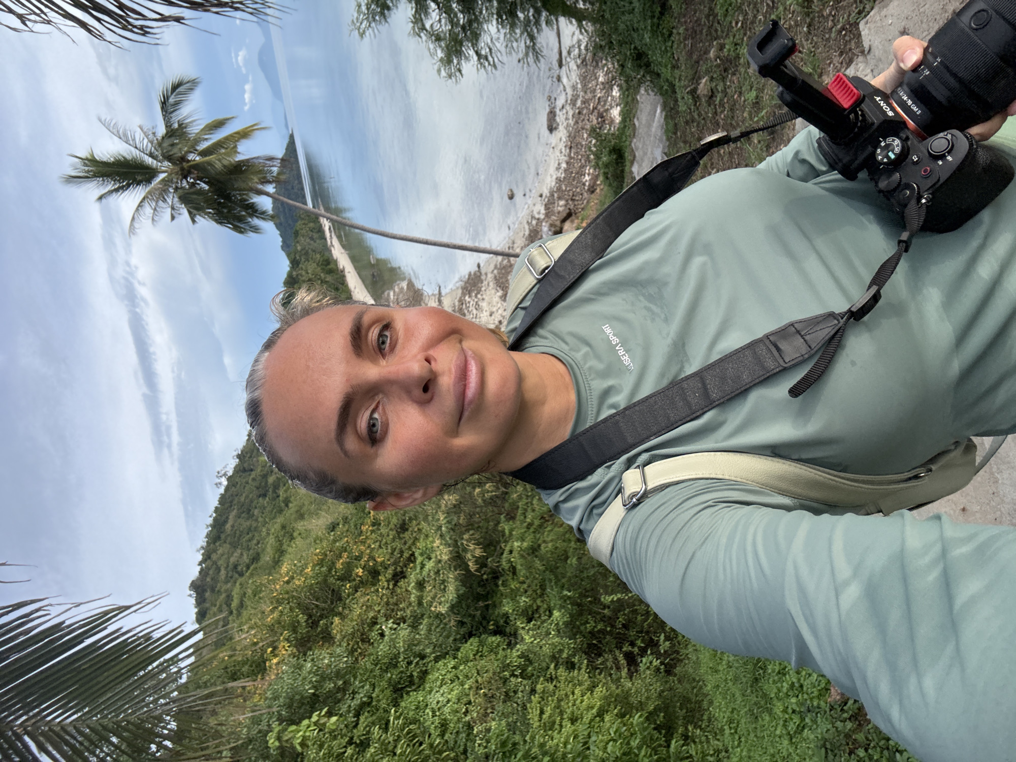 In the foreground, a woman with blonde hair tied back, facing forward, takes a selfie. She is wearing a long-sleeved, light green t-shirt with "USERA SPORT" visible on the left chest. A black camera strap crosses her chest, and a light-colored bag is slung over her right shoulder. She holds a black Sony camera with a lens in her right hand. Her skin appears tanned with a slight sheen.
The background reveals a tropical landscape. Lush green foliage covers a hillside to the left, with a clear blue sky above. To the right, a calm body of water, possibly the sea, meets a sandy shoreline dotted with debris. A prominent palm tree leans over the water, its fronds extending towards the top left of the frame. In the distance, hills rise from the water under a cloudy sky. The scene is consistent with a coastal area in Baan Pang Ka, Thailand.