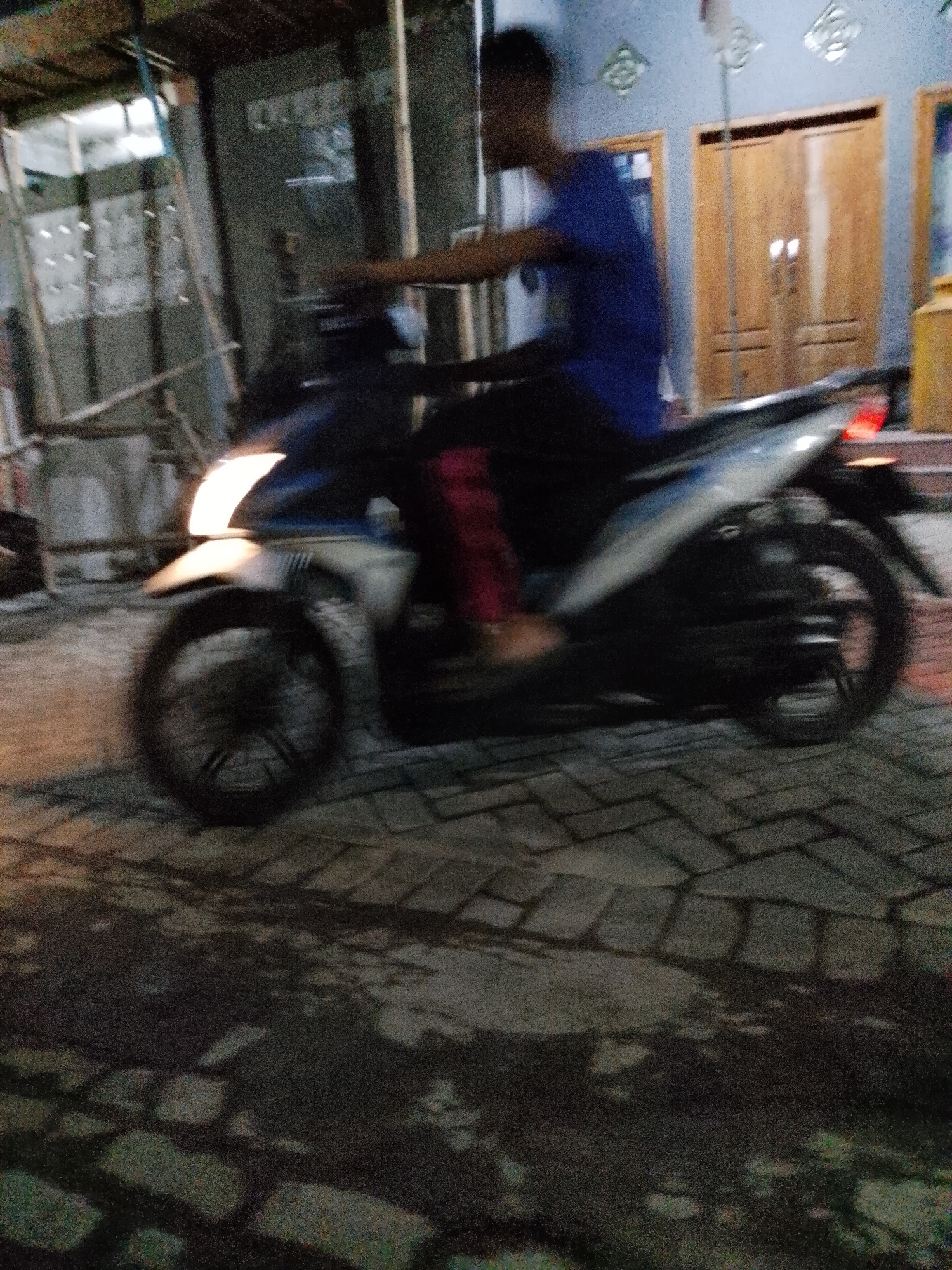 A motion-blurred image depicts a person on a scooter at night on a brick-paved road in Pasuruan, Indonesia. The person, wearing a blue t-shirt and dark pants, is positioned on a scooter with a white and blue body. The scooter's headlight is illuminated, casting a bright light. In the background, there is a building with wooden doors. The overall scene is dimly lit, with shadows and motion blur obscuring fine details.