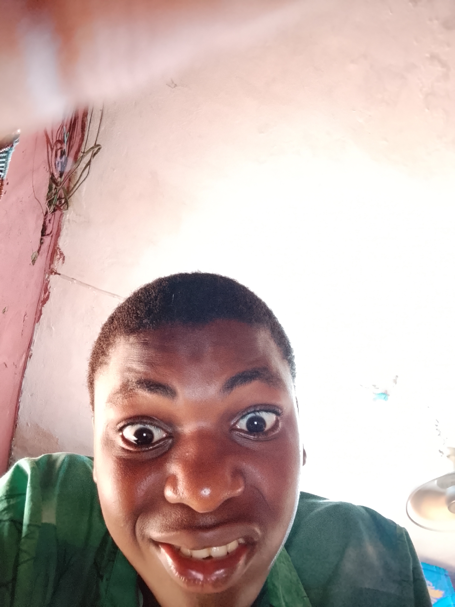 A low-angle, close-up photograph features a person's face, appearing to be male, looking directly into the camera. The individual has dark skin, short dark hair, and is wearing a green collared shirt. They exhibit an exaggerated, playful expression with widely opened eyes, dilated pupils, and an open mouth revealing teeth in a broad grin.
The background is brightly lit, almost overexposed in the upper sections. A light pink or peach-colored wall is visible on the left, with several entangled wires or cables running along it. On the far right, a portion of a light fixture, possibly a ceiling fan, is discernible. The setting appears to be an indoor room, characterized by simple wall textures and visible electrical wiring. The lighting suggests either bright daylight or strong artificial illumination. No text is visible in the image.