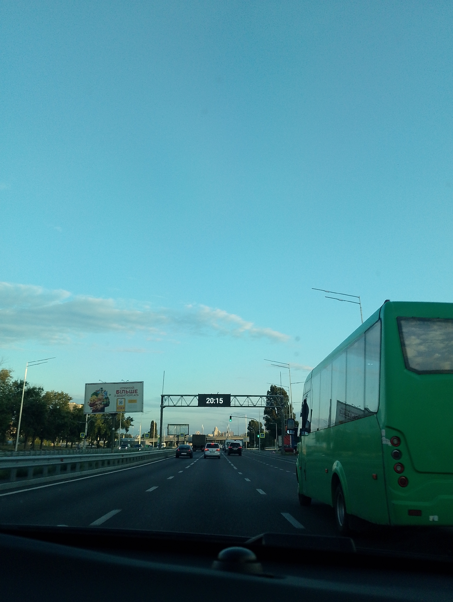 The image shows a view from inside a vehicle on a highway in Kyiv, Ukraine, as indicated by the billboard text and the digital display showing 20:15. A green bus is prominently featured on the right side of the frame, traveling in the same direction as the viewer's vehicle. The highway has multiple lanes, and other vehicles are visible in the distance. To the left, there is a billboard with Ukrainian text and an advertisement, along with trees and streetlights. The sky is blue with some scattered clouds, suggesting it is either early morning or late afternoon. The overall scene depicts traffic on a major road within the city.