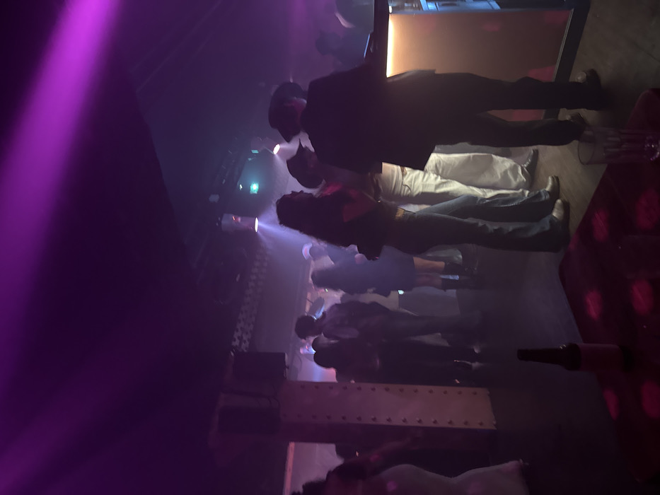 The scene shows a crowded nightclub with dimmed lights and purple spotlights. People are standing and dancing, their silhouettes highlighted by the lighting. A man in a dark suit stands near a bar or counter. The atmosphere suggests a lively, social gathering with a focus on entertainment and nightlife. A bottle is visible in the foreground.