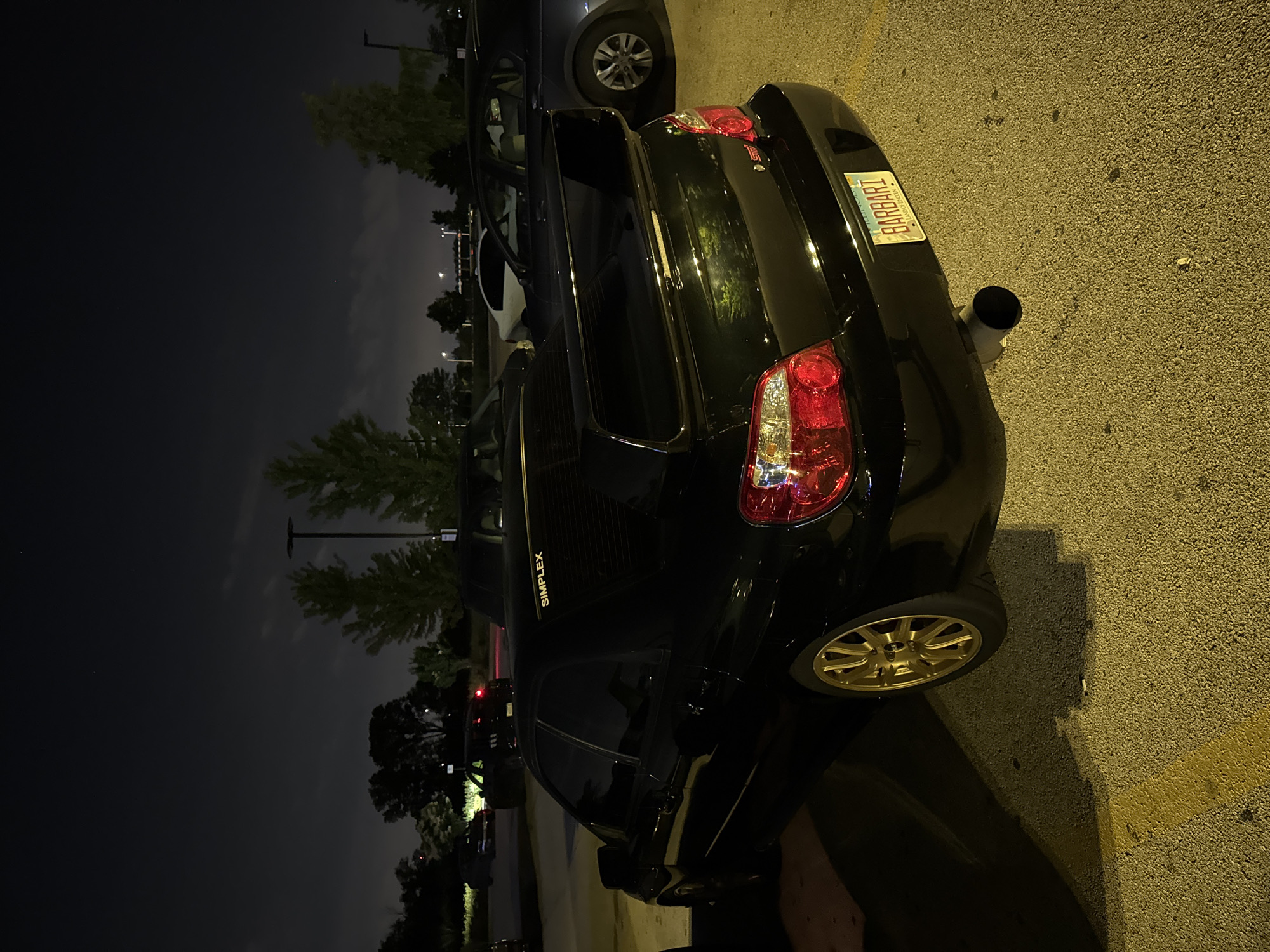 In the city of Schaumburg, United States of America, a black Subaru WRX STI is parked in a dimly lit outdoor parking lot at night. The car, viewed from its rear three-quarter angle, exhibits a large rear wing, gold multi-spoke rims, and a visible exhaust pipe. The license plate reads "BARBARI" and is bordered by a yellowish frame with the words "LAND OF LINCOLN" printed below. Above the rear window, a decal reads "SIMPLEX". The parking lot's asphalt surface is marked with a yellow line. The background features trees and streetlights, suggesting an urban or suburban setting.