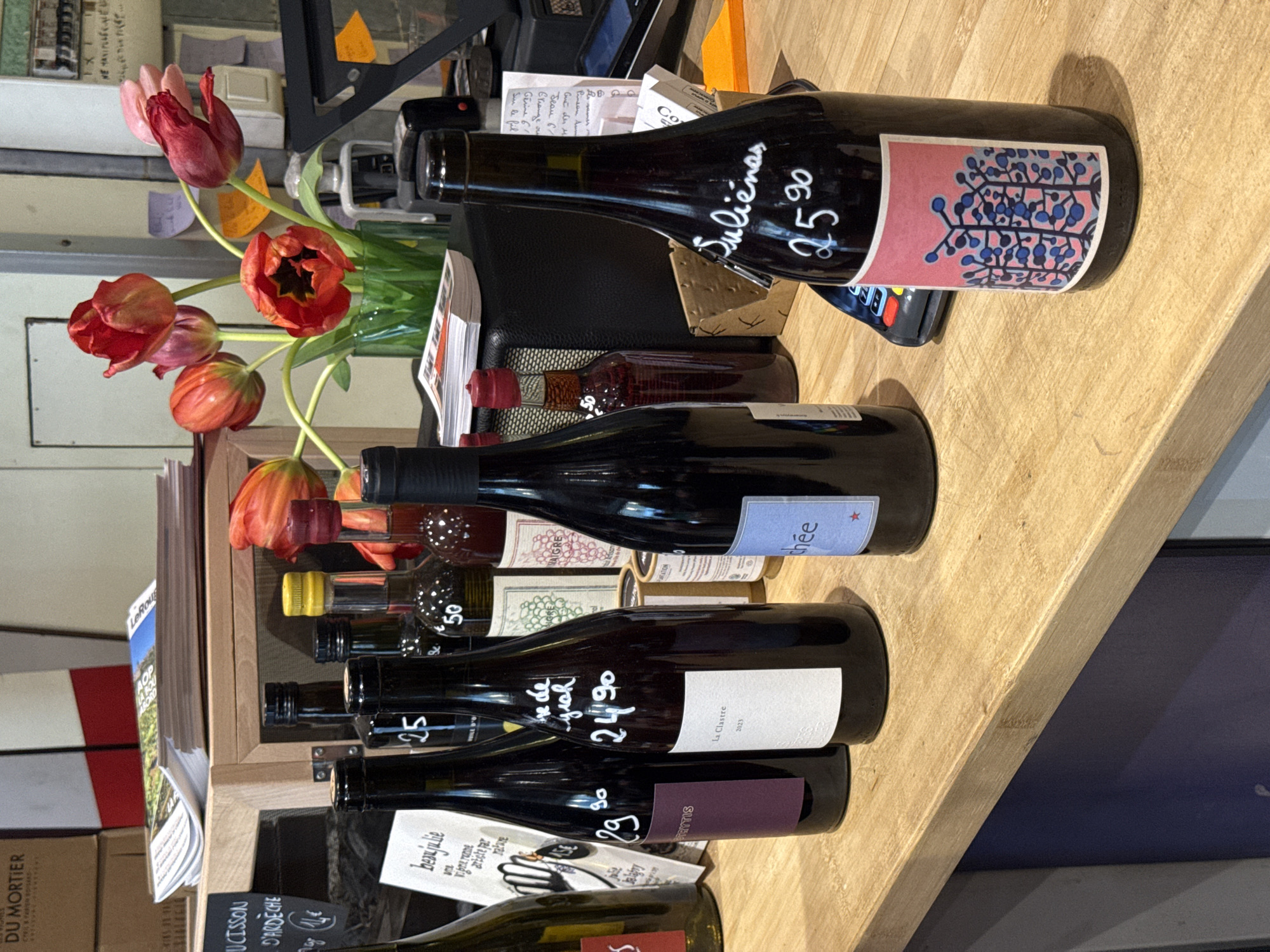 A still-life photograph shows a selection of wine bottles arranged on a light wooden surface, likely a counter or shelf, possibly in a store or shop. The scene includes several bottles of red wine, with prices written on them in white ink, such as "Julienas 25.90". Various labels are visible, including "La Clastre 2023" and "beanjulie." Tulips in a vase add a touch of color to the display. The background includes boxes and printed materials, suggesting a commercial environment.