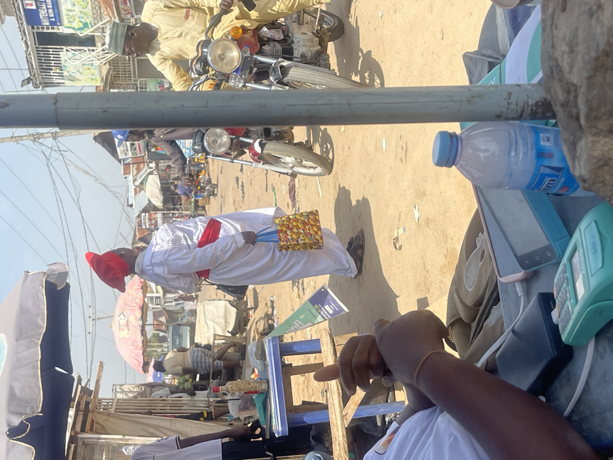 The image depicts a street scene in Abuja, Nigeria, possibly a market or a busy thoroughfare. In the foreground, a person's arms and hands are visible, resting on a surface with electronic devices, including a black power bank, a pink smartphone, and a green electronic device with a keypad. A bottle of water with a blue cap and a partially visible label is also present. In the middle ground, a person dressed in white with a red sash and a red headdress is holding a patterned bag. Behind this person, two motorcycles are parked, with riders visible on them. The environment is dusty with sparse vegetation and various stalls or buildings lining the street, indicating an outdoor commercial area. Power lines are visible overhead.