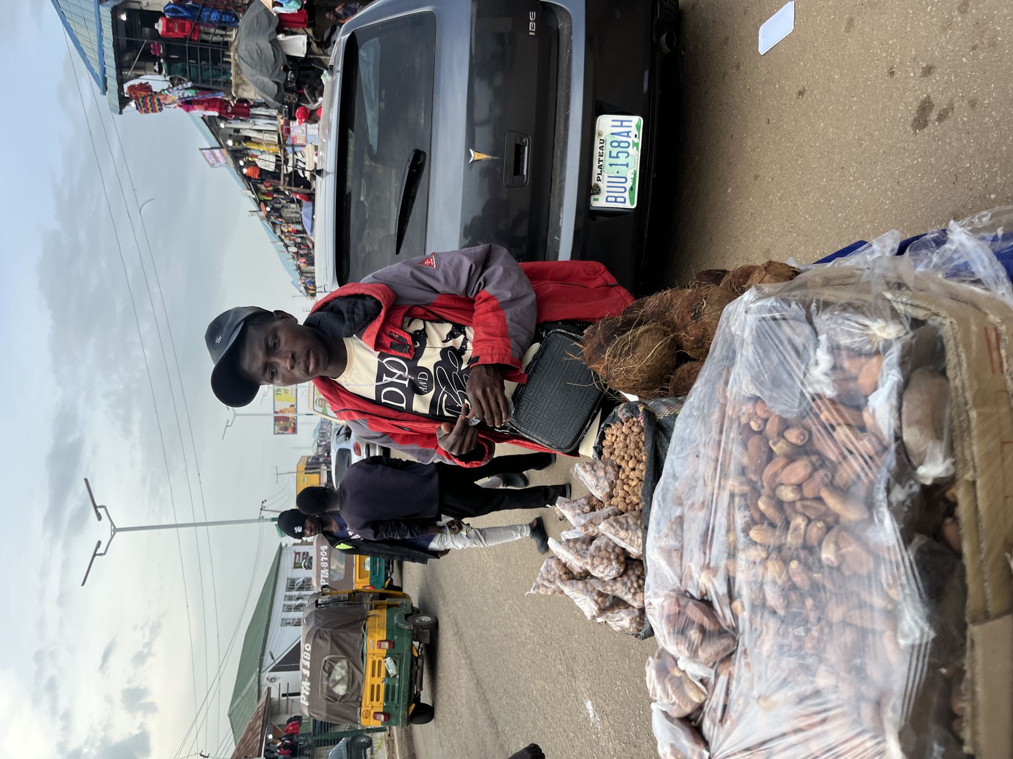 In Bukuru, Nigeria, a bustling street market scene unfolds. The central subject is a young man, likely a vendor, dressed in a red and grey jacket over a cream-colored t-shirt, and wearing a black baseball cap. He is positioned near a black SUV, its rear visible, bearing a license plate that reads "PLATEAU BUU-158AH". Next to him are piles of produce, including coconuts and numerous small, bagged items that appear to be nuts or dried fruits.
The setting is an outdoor market with stalls lining the street, filled with various goods, including clothing displayed on racks. Several other people are present, some appearing to be vendors or customers, moving through the market. A yellow and green auto-rickshaw, a common mode of transport in the region, is parked nearby, with visible text on its side, including "PTA 8704" and "PTA-1186". The ground is paved with asphalt.
The time of day appears to be daytime, with an overcast sky suggesting a possibly cloudy or slightly overcast weather. The overall atmosphere suggests a lively commercial activity, typical of a Nigerian market. The expressions of the people are not clearly discernible to infer specific emotions, but the presence of goods for sale and the movement of people indicate ongoing transactions and daily commerce.