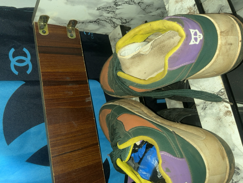 The scene depicts a pair of multi-colored athletic shoes resting on a dirty, light-colored surface with a mottled, marble-like pattern. The shoes are primarily dark green with orange panels, yellow ankle trim, and purple heel counters. The right shoe's heel features a white, stylized fox head logo. Dark green laces are present, with one untied and draped. The soles are thick, light tan, and appear dirty. The interior of the left shoe contains blue plastic material, while the right shoe's interior lining is visible and soiled. Above the shoes, a horizontal wooden panel with a brown grain is present, secured on the right by two brass-colored metal brackets. Behind and above this panel, a dark blue or black fabric is visible, displaying light blue graphics, including a partially obscured interlocking "CC" logo. Other dark, indistinct objects are partially visible beneath the shoes. This scene is located in Ilishan, Nigeria. No people are visible.