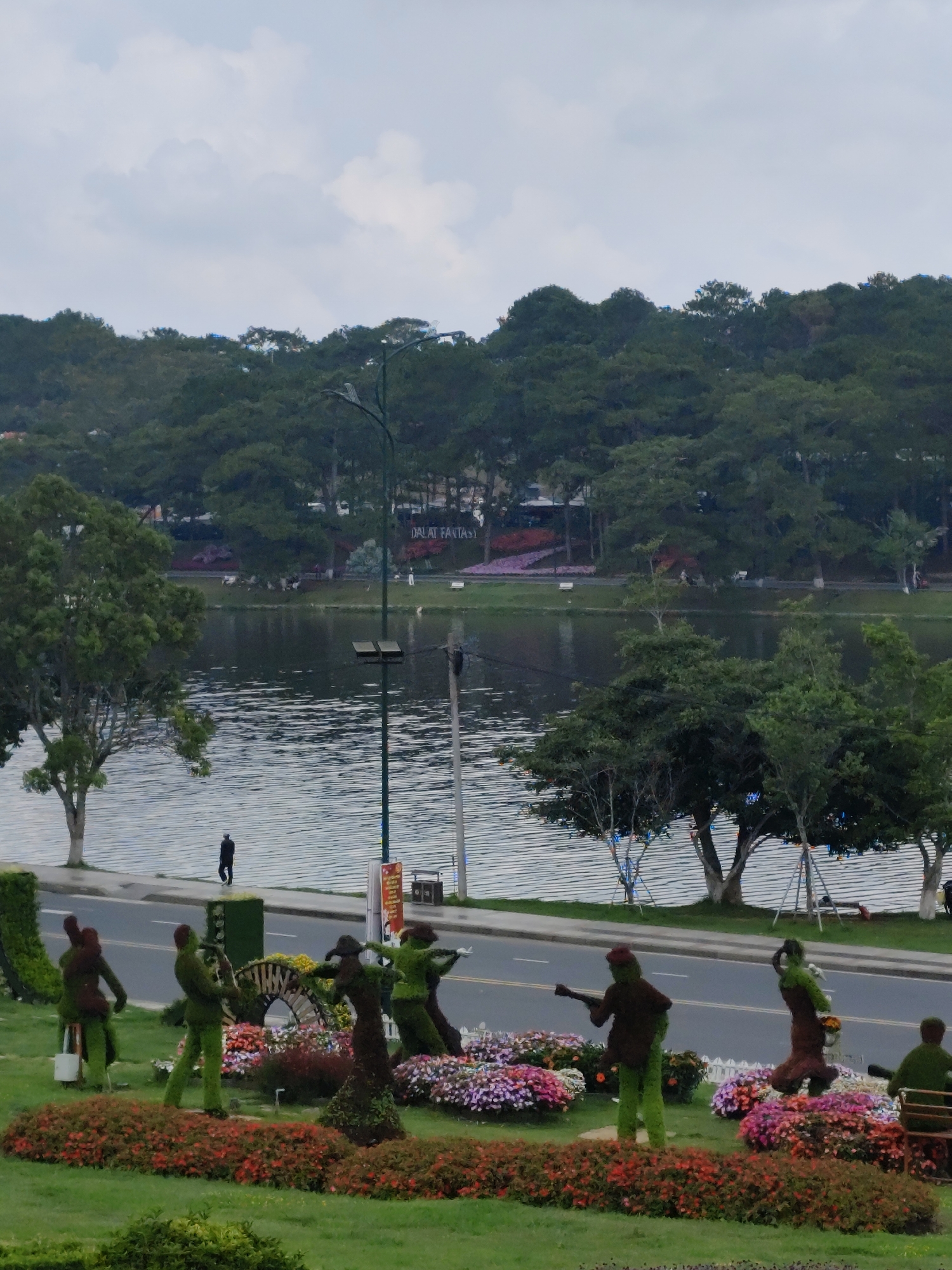 This image captures a vibrant and serene outdoor scene in Da Lat, Vietnam, characterized by a mix of natural elements and decorative landscaping under an overcast sky.
In the foreground, a meticulously designed garden display features several topiary figures crafted from green foliage and brown accents, arranged to depict a musical ensemble. One figure appears to be playing a guitar, another a saxophone-like instrument, a third is seated, and another strikes a dynamic pose as if dancing or singing. These topiaries are surrounded by lush flower beds bursting with red, pink, purple, and yellow blossoms, along with manicured green grass.
A paved road separates the garden display from a large, tranquil lake that dominates the midground. The lake's surface shows gentle ripples, reflecting the cloudy sky and surrounding greenery. On the far side of the road, a lone individual in dark clothing walks along the lakeshore. A tall street lamp or utility pole stands prominently beside the road.
The background is dominated by a dense forest of pine trees covering rolling hills, stretching along the far bank of the lake. Amidst the trees on the far bank, a white sign with black lettering clearly reads "DALAT FANTASY," confirming the location context. Other distant flower beds and paths are also visible on the far side.
The overall impression is one of a peaceful, well-maintained park or scenic area, likely a tourist attraction given the decorative elements and the sign. The overcast weather casts a soft, diffused light over the entire landscape.
