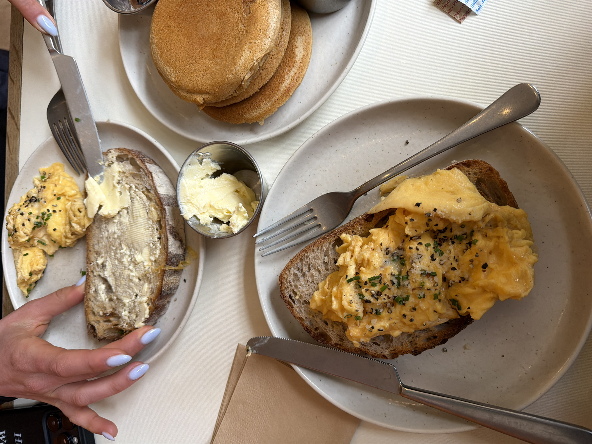 A high-angle shot shows a table with two plates of breakfast food and a stack of pancakes, likely in a cafe in London, UK. On the left, a woman’s hand with light blue manicured nails holds a fork and knife, spreading butter on a slice of bread served with scrambled eggs on a white plate. A small metal container of butter is also present. A stack of pancakes sits on a separate white plate. On the right, a second plate contains another slice of bread topped with scrambled eggs, accompanied by a fork and knife. A brown napkin and a mobile phone are visible at the bottom of the frame. The table has a white textured surface.