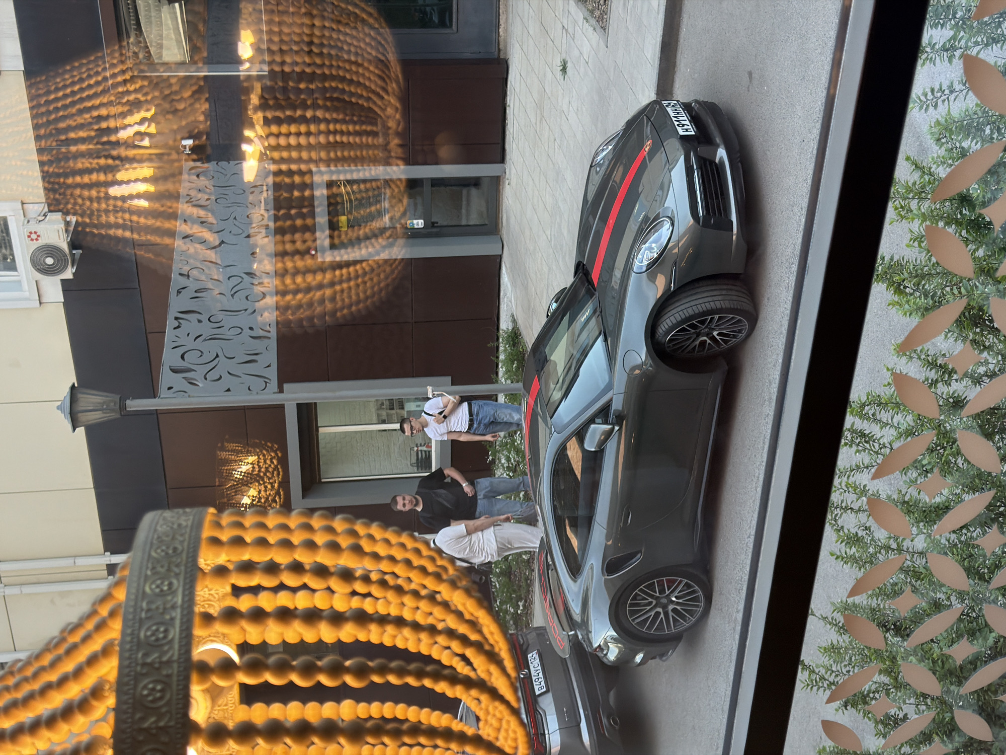 The image, taken in Krasnoyarsk, Russia, captures a street scene viewed through a reflective window. In the foreground, a large, ornate, beaded light fixture, resembling a chandelier, dominates the left side, casting warm, circular patterns of light onto the building facade opposite.
On the street, two dark-colored cars are parked along the sidewalk. The prominent vehicle is a dark grey or black Porsche, likely a 911 model, distinguished by a red racing stripe extending down its hood and roof. It is equipped with multi-spoke dark alloy wheels. Behind it, a second dark car is partially visible. Two Russian license plates are identifiable: "H971HH186" on the front car and "B494YC185" on the partially visible rear car.
Three men are standing on the sidewalk near the cars. One man wears a white t-shirt, dark pants, and glasses. Another is in a black t-shirt and light blue jeans. The third, partially obscured by the foreground light fixture, also appears to be in a white t-shirt and light-colored pants. They appear to be casually gathered.
The background shows a modern building facade with dark panels and a light-colored decorative metal screen featuring intricate, swirling patterns. A street lamp and a white air conditioning unit are also attached to the building. On the far right, green artificial foliage with cut-out floral patterns is visible, suggesting another decorative element or screen. The lighting suggests it is late afternoon or early evening, characterized by warm artificial light and natural ambient light.