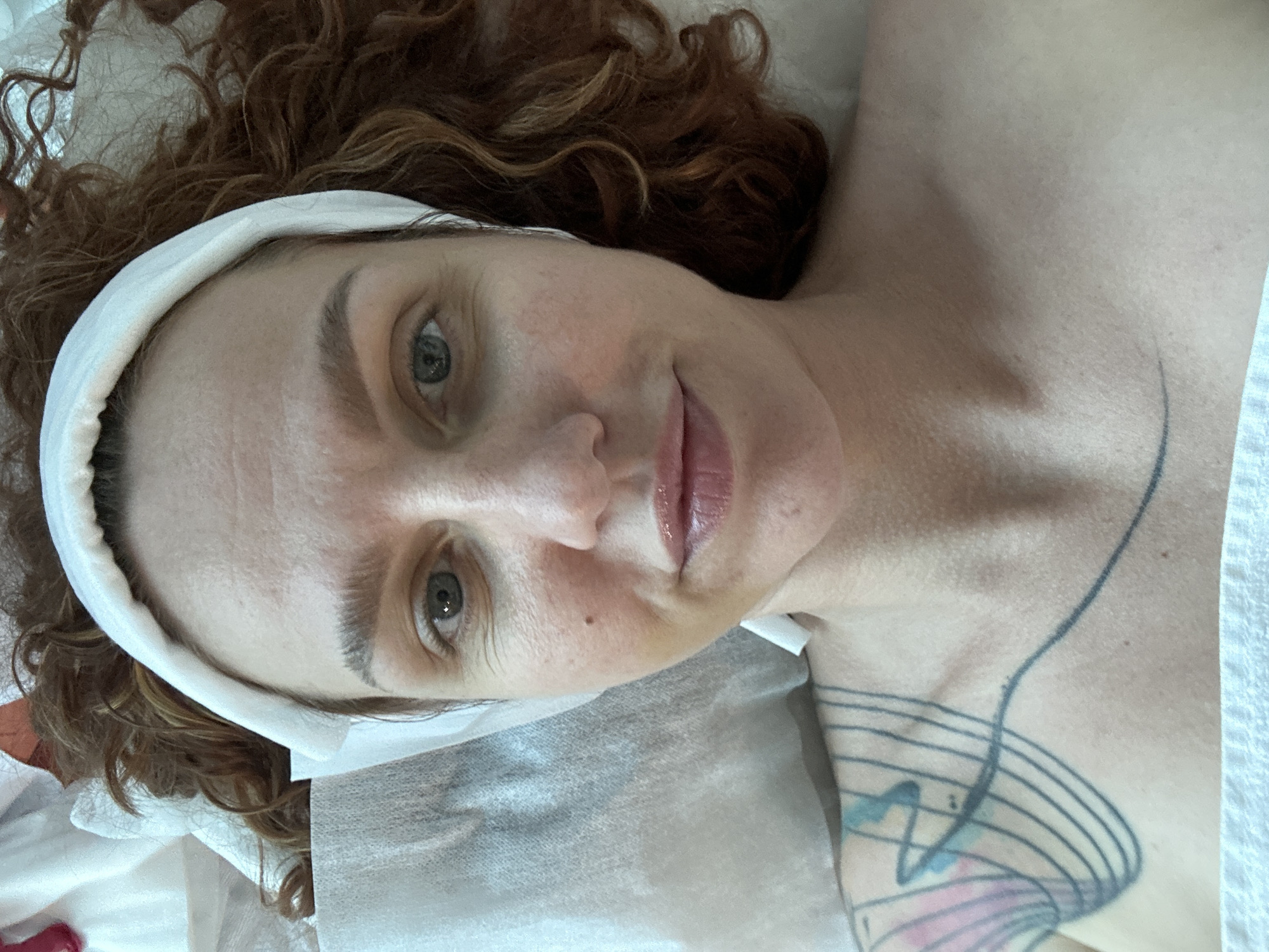 A light-skinned woman with curly reddish-brown hair, wearing a white headband, lies on a bed covered with a gray sheet. Her eyes are open, and she has a slight smile. A tattoo of musical notes and a design can be seen on her upper chest and shoulder. The background shows a blurred room. No evidence suggests this image was taken in Sochi, Russia.