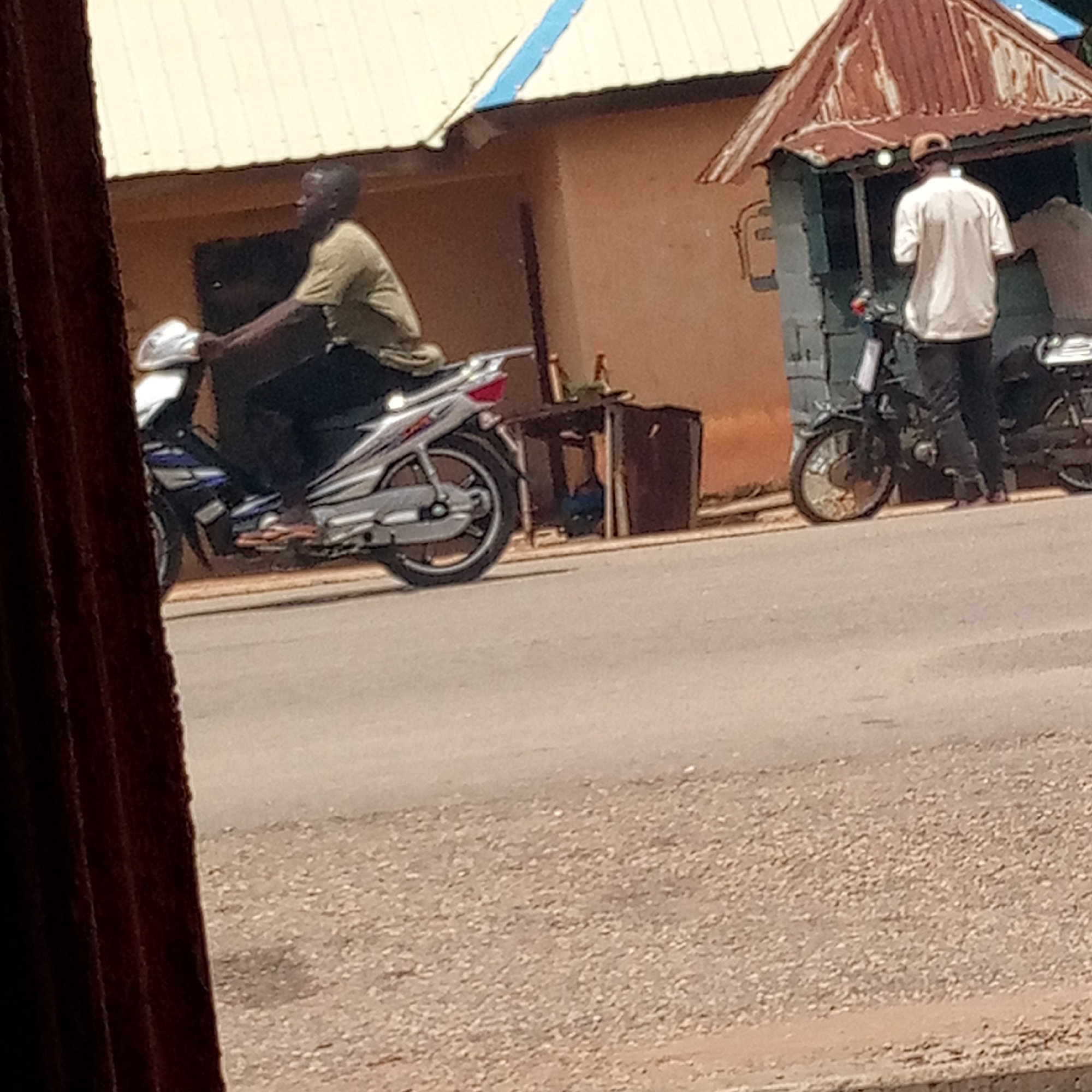 The image depicts a street scene in Mararaba, Nigeria. A man is riding a silver and blue motorcycle on the road. To the right, a man stands near a small, open-fronted structure with a rusted corrugated metal roof. There are two motorcycles parked near the structure. A small table with items on it sits between the man on the motorcycle and the structure. The road appears to be paved but somewhat worn. The buildings in the background are of simple construction. A section of a door frame is visible in the foreground, suggesting the image was taken from inside a building.