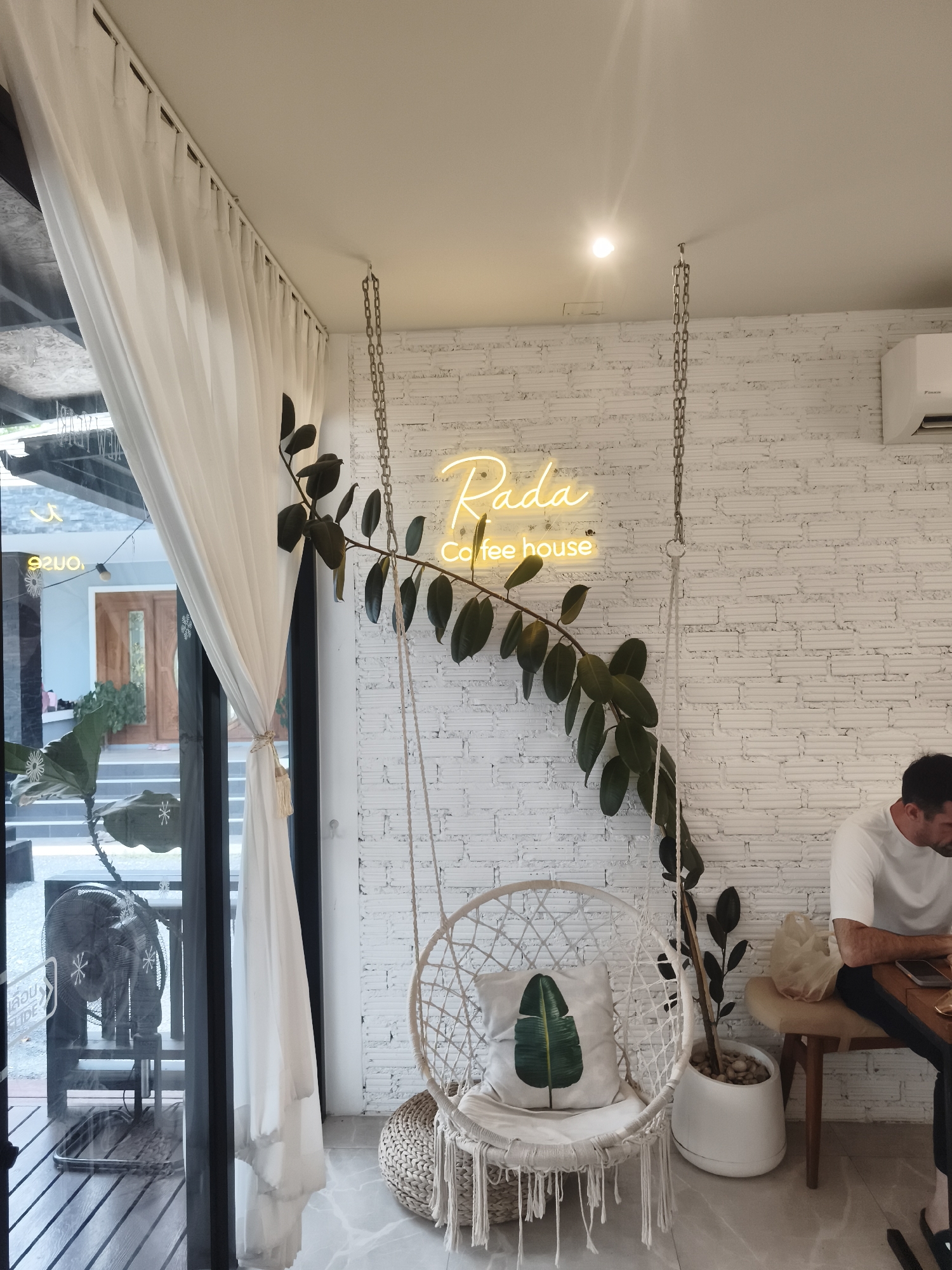 The image shows the interior of a coffee house. In the foreground, a white macrame hanging chair with a leaf-patterned cushion is visible, placed on a woven mat. To the right, a potted plant in a white planter sits on the floor. A man in a white t-shirt is seated at a table to the right, looking down at his phone. In the background, a white brick wall is adorned with a neon sign that reads "Rada Coffee House". A white curtain hangs on the left side of the frame, partially obscuring a view of the outdoors, which includes a doorway and some plants. An air conditioning unit is mounted high on the wall. The overall ambiance suggests a modern and relaxed cafe setting in Ko Samui District, Thailand.