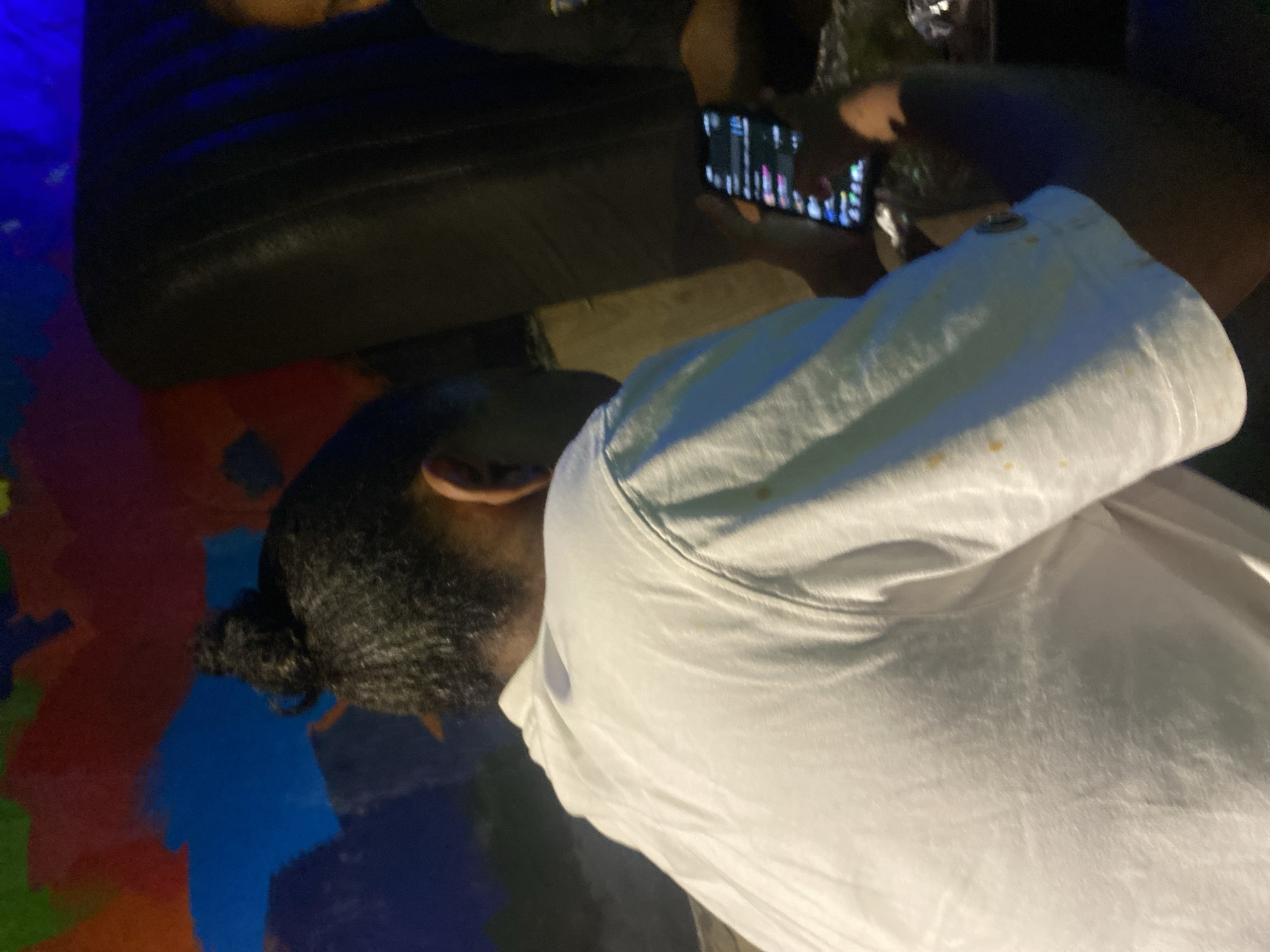 The image shows a person from behind, wearing a white t-shirt with some stains, and their hair styled in a bun. They are holding a smartphone, which displays a list of items. The person is seated in what appears to be a dimly lit indoor setting with a dark, possibly leather, couch visible to the right. Behind them, a wall is decorated with a colorful, abstract mural. Another person's shoulder and head are partially visible on the right side of the image, also in shadow. The overall ambiance suggests a casual gathering or a relaxed environment. While the location is stated as Abuja, Nigeria, there are no specific visual cues within the image to confirm this location.