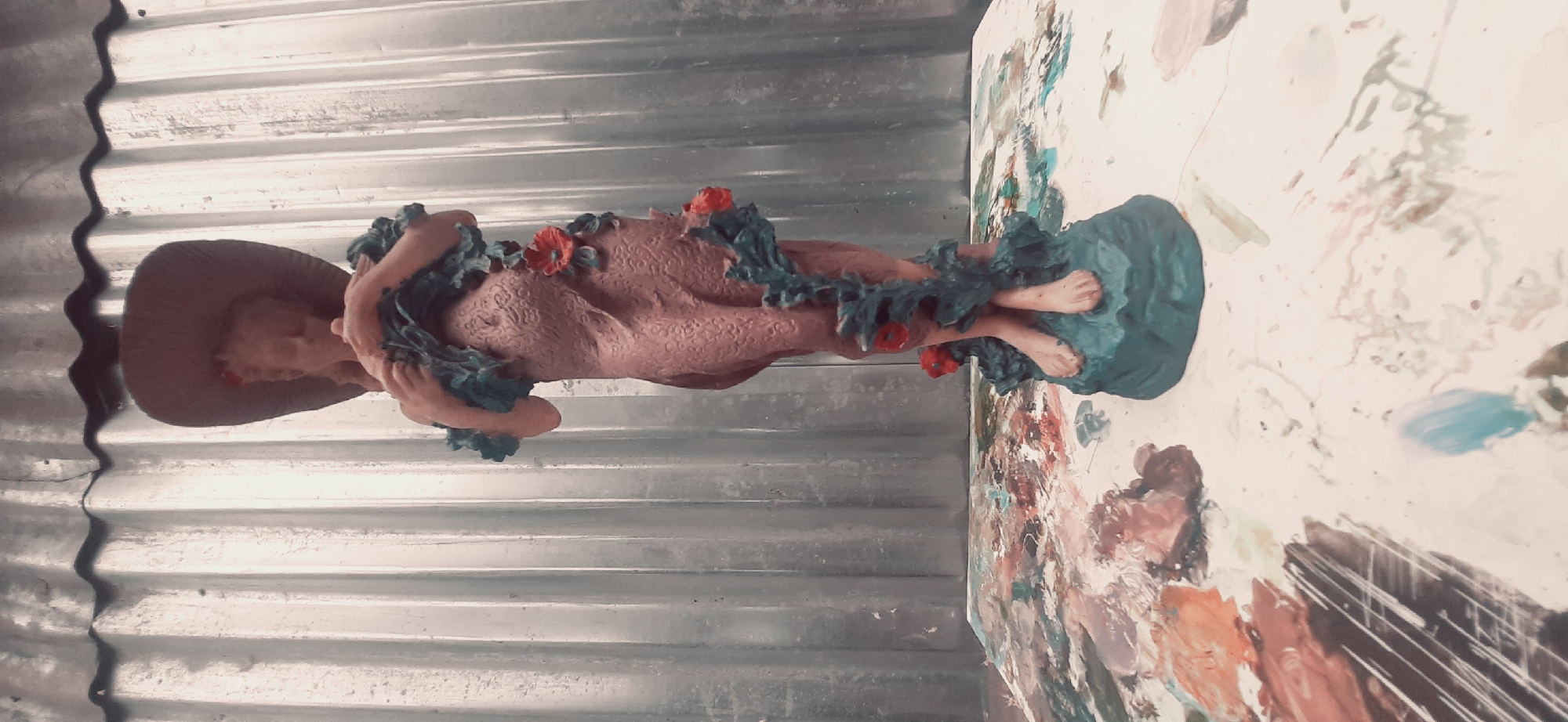 A full-body statue of a woman with vines and red flowers wrapped around her is displayed on a palette covered in colorful paint splatters. The statue is positioned in front of a corrugated metal wall. The woman is wearing a wide-brimmed hat and her eyes are closed. She is holding something to her chest. The statue stands on a teal-colored base, shaped like a rocky outcrop. The palette appears to be made of plastic and has various shades of paint, including orange, brown, white, and blue, applied in a messy, artistic fashion. The background wall is a light gray color and has vertical corrugations. The overall lighting suggests an indoor setting, possibly a studio or workshop. There are no visible people or any indication of the specific location within Yessentukskaya, Russia.