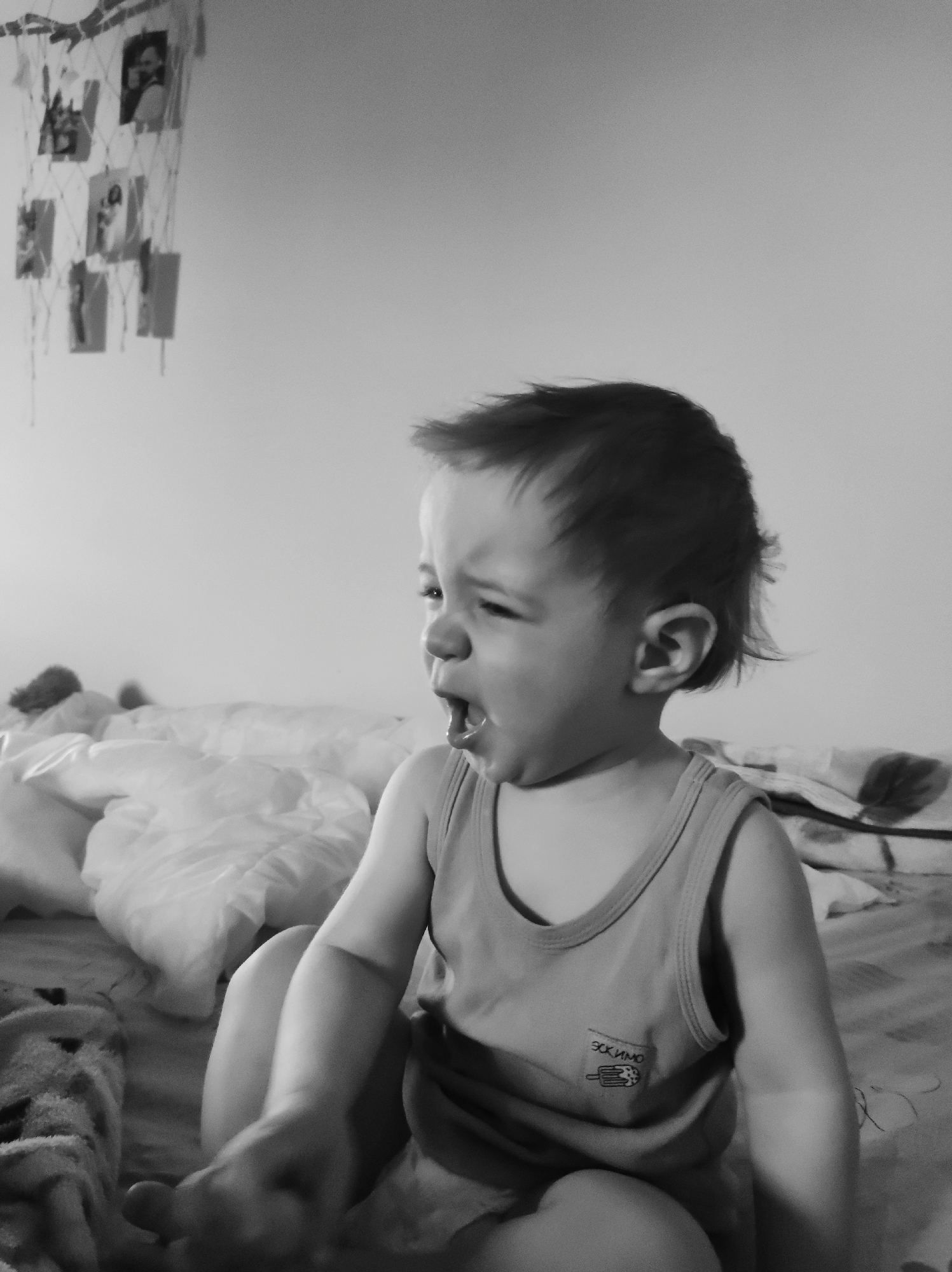 A black and white medium shot depicts a young child sitting on a bed in a room. The child has short hair and is wearing a tank top with a small design on the upper left side. The child's mouth is open in what appears to be a yell or cry, with their head turned to the side. The bed is covered with a wrinkled blanket and a plush, crumpled pillow. In the background, against the wall, there's a decorative item resembling a net with photos attached. The location is Unknown, Unknown.