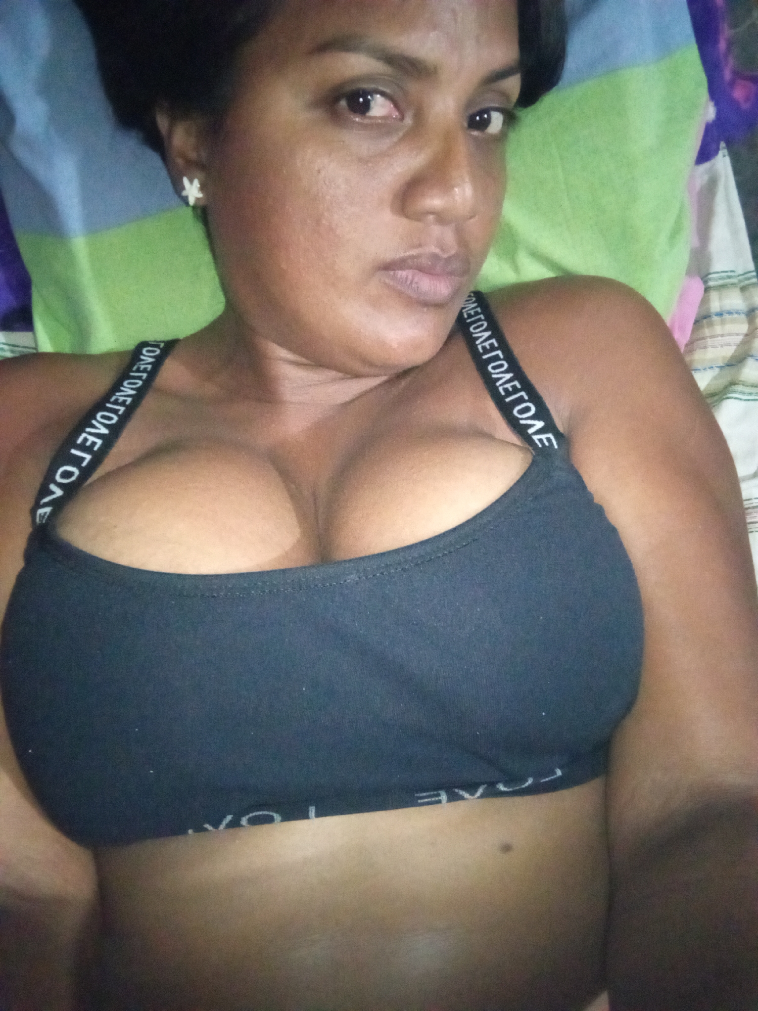 A close-up, eye-level indoor shot depicts a person, likely female, with dark skin, dark hair, and dark eyes, lying down. She looks directly at the camera with a neutral expression. She is wearing a black sports bra or bralette, the straps of which feature white repeated text, visibly spelling "LOVE" (or "EVOL" when seen in reverse due to perspective). A white, star-shaped earring is visible on her left ear. The background behind her consists of multi-colored pillows or bedding with striped patterns in shades of green, blue, and off-white. The lighting is soft, suggesting an indoor setting, possibly a bedroom, where the person is posing for a photograph.
Location context: Unknown, Unknown