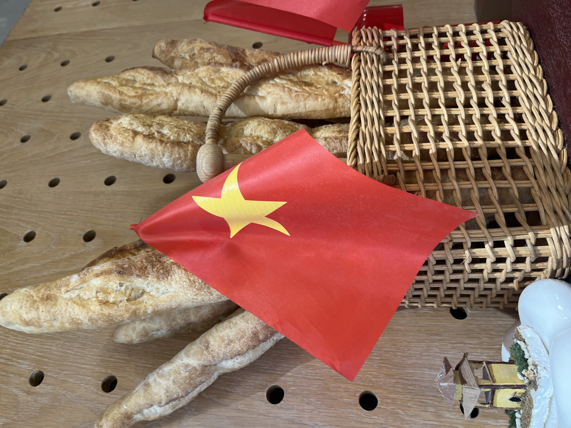 The image shows a scene with several objects arranged on a light brown wooden surface. There are about seven crusty baguettes stacked near the center and left side of the frame. Next to them is a woven basket with a handle, containing more baguettes. A red flag with a yellow star is placed prominently in front, partially obscuring the bread. To the bottom right, a small decorative house or figurine is visible. The setting appears to be indoors, possibly on a table or countertop. The location is not identifiable from the image.