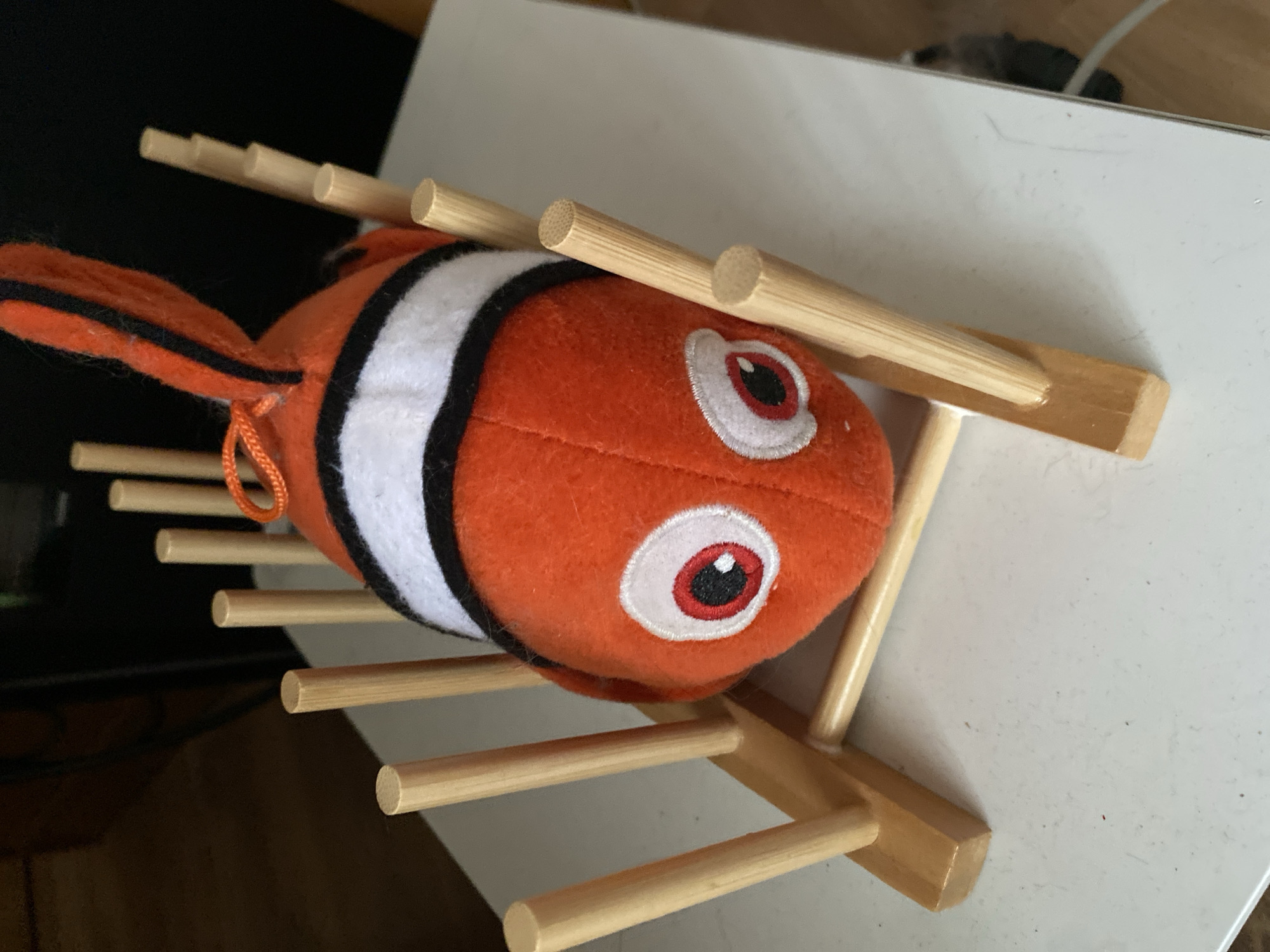 An orange stuffed clownfish with white and black stripes is placed in a wooden holder. The holder is made of several vertical wooden dowels connected by horizontal wooden pieces. The clownfish has large, googly eyes with red and black irises and white pupils. It is positioned head-first in the holder, with its tail fin extending upwards. The holder and clownfish are resting on a white surface, possibly a table, with a blurred background that suggests an indoor environment. The lighting is overhead and somewhat diffuse. There is no indication of the location being in Bílina, Czechia.