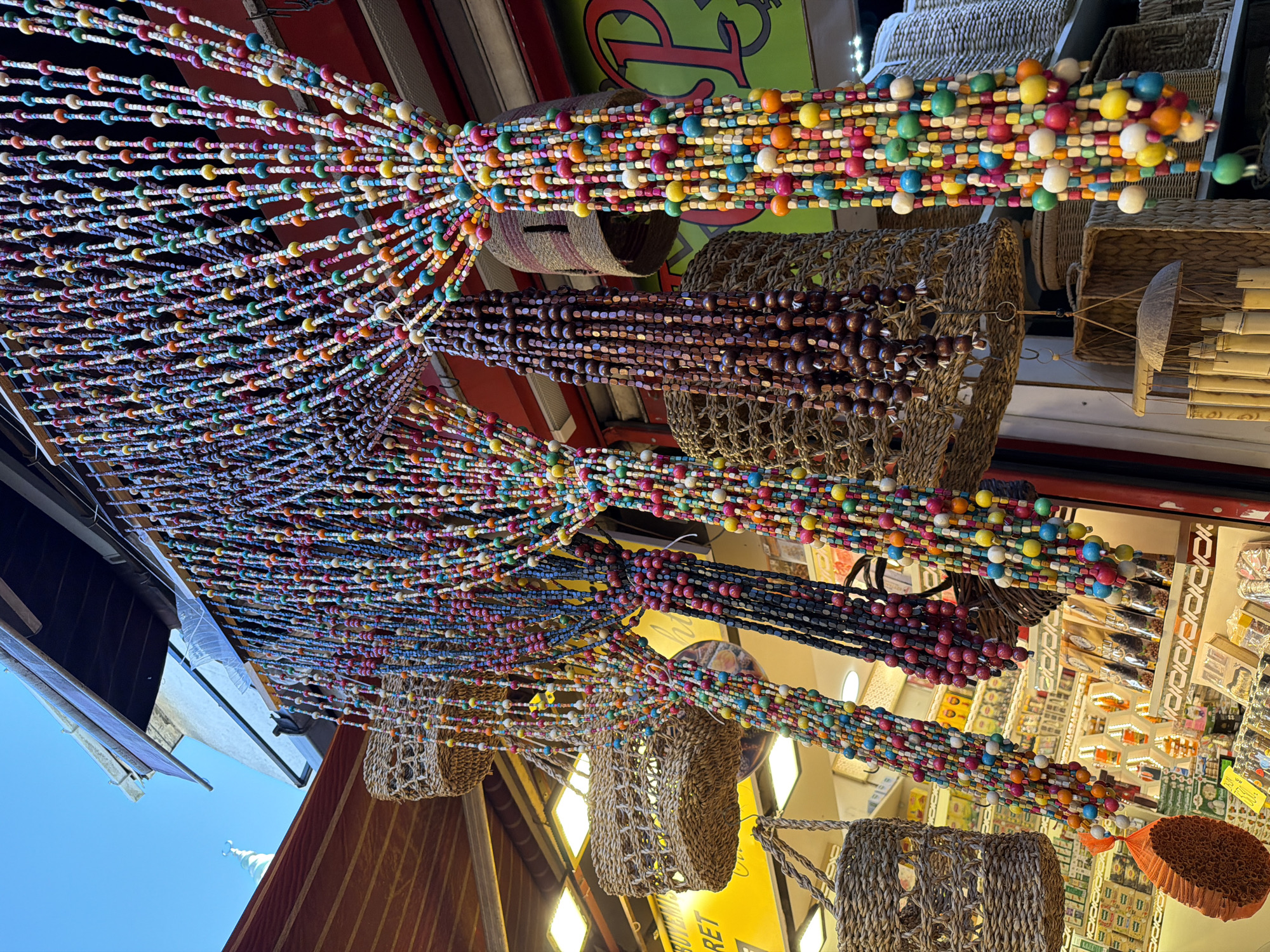 The image shows a street market scene in Istanbul, Turkey, with a focus on colorful beaded curtains and woven baskets. The beaded curtains are made of many strands of small, round, and square beads in a variety of bright colors, including red, blue, yellow, green, and white. These curtains hang in front of a shop with shelves displaying various goods. Several woven baskets are also visible, some hanging and others placed on shelves. The baskets appear to be made from natural materials like straw or wicker and have intricate weaving patterns. The background shows a shop interior with warm lighting and some signage that is partially visible, with a green sign with a red "P" being the most prominent. Above the shop, awnings and a clear blue sky are visible, suggesting an outdoor setting in daylight. The overall impression is one of a vibrant and artisanal market, likely a tourist area.