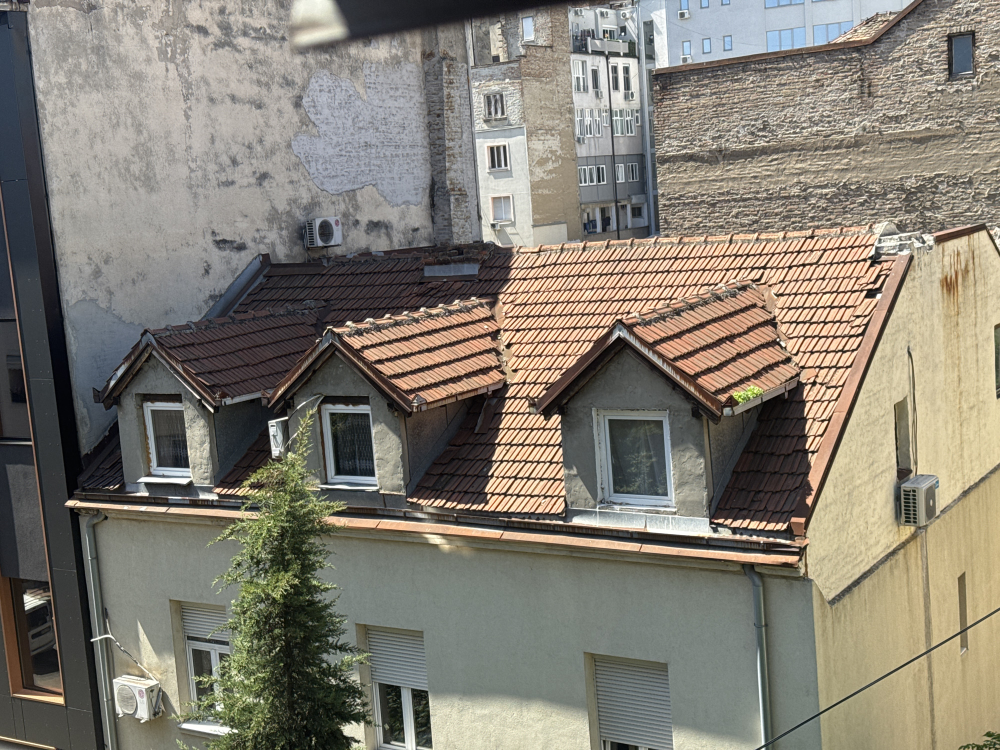 The image shows a view from above, looking down at rooftops and the facades of several buildings in Zvezdara Urban Municipality, Serbia. The foreground features the tiled red roof of a light-colored building with several dormer windows. A tall, slender evergreen tree is positioned in front of this building, partially obscuring the lower floors. To the left, a modern building with a dark facade and reflective windows is visible. Behind these buildings, older, multi-story structures are seen, characterized by varying architectural styles, weathered facades, and numerous windows. Some buildings display exposed brickwork and signs of age. Several air conditioning units are mounted on the exterior walls of the buildings. The scene is bathed in daylight, casting shadows and highlighting the textures of the building materials.