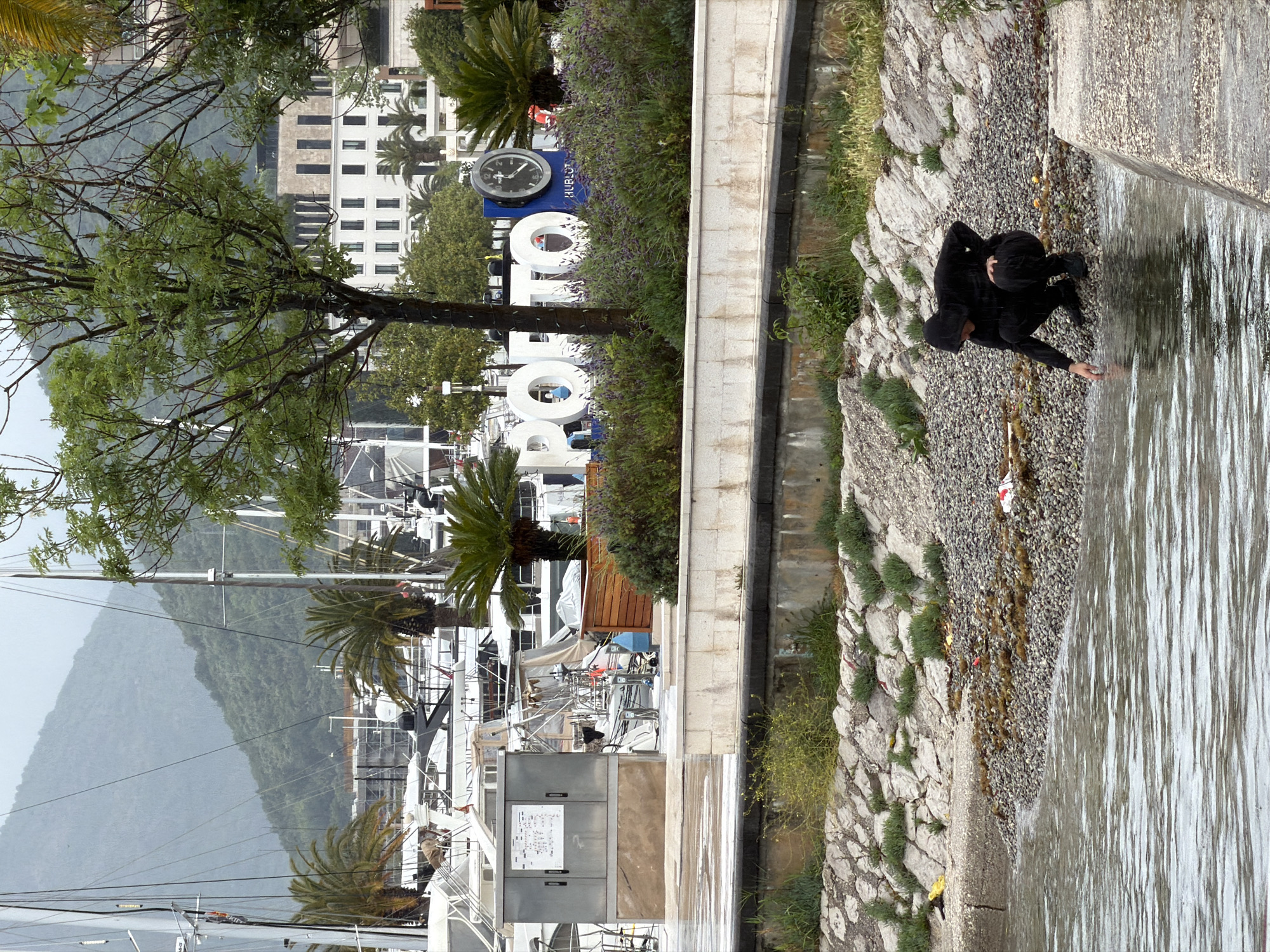 The image captures a scene in Tivat, Montenegro, featuring a person dressed in black, likely a hooded jacket and pants, standing near the edge of a body of water, potentially the Adriatic Sea, with their hand reaching towards the water. The shore is pebbly and rocky, with some vegetation interspersed.
In the background, there is a harbor with numerous yachts and sailboats docked, their masts visible. Further inland, a tree with bare branches partially obscures a view of a building with multiple windows. Next to the building, there is a display with a large clock and the word "PORTO" spelled out in capital letters. The landscape transitions into a mountainous terrain in the distant background. The lighting suggests it is daytime with overcast or diffused light.
