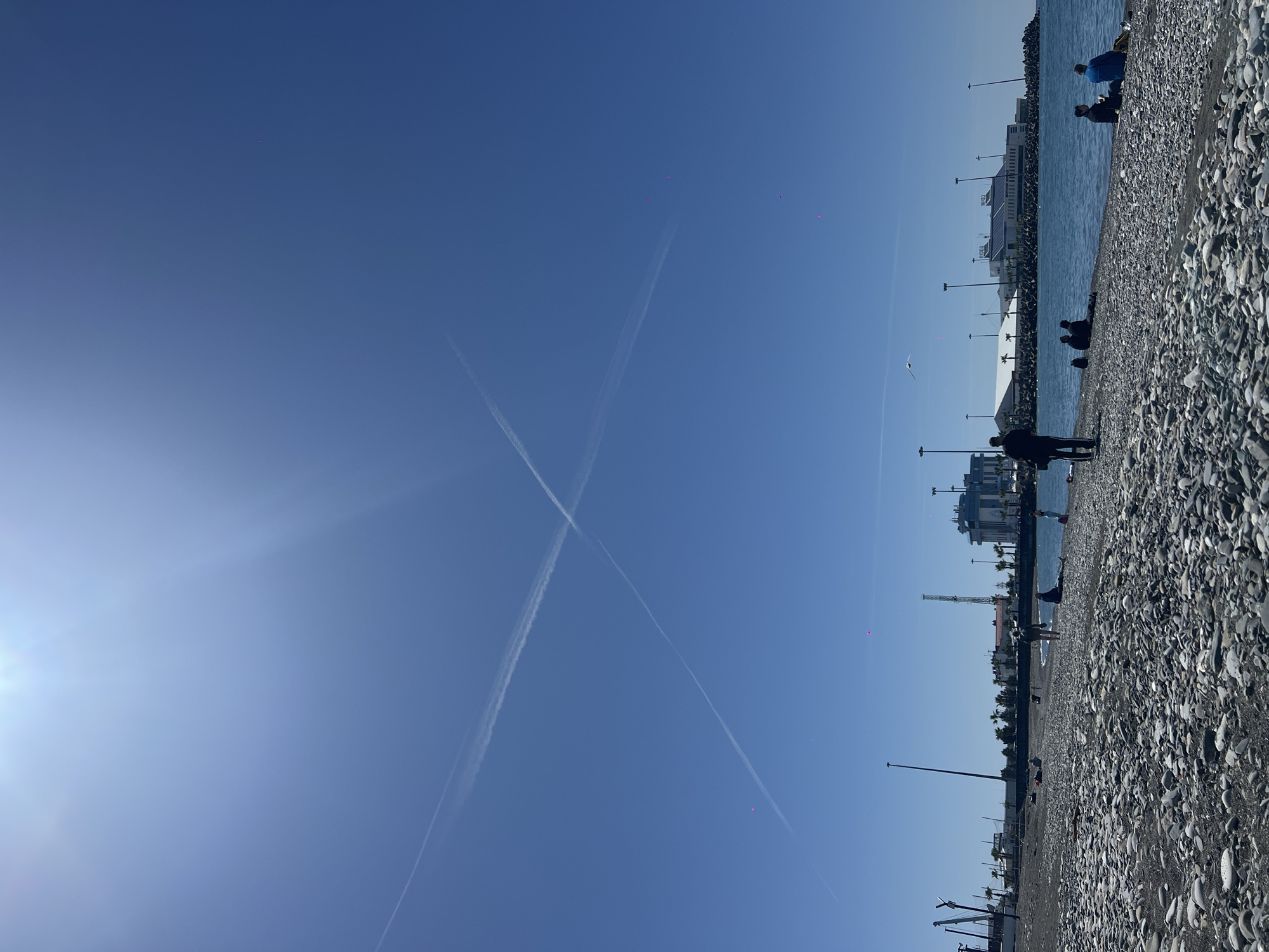 The image captures a bright, sunny day at a pebble beach in Sochi, Russia. The sky is a clear blue, dominated by two prominent airplane contrails that cross to form an "X" shape. A strong sunburst is visible on the left side of the frame. Several small, distinct magenta specks are scattered across the sky, which may be image artifacts or tiny distant objects.
Along the right side of the image, a wide pebble beach stretches towards calm, blue water, likely the Black Sea. Several people are present; one individual in a dark jacket stands on the pebbles, while others are seated or positioned closer to the water's edge. In the background, across the water, a series of modern buildings line the shore. One multi-story building, equipped with solar panels on its roof, clearly displays the Cyrillic text "СОЧИ" (SOCHI). Other structures, possibly part of a port or marina, along with lamp posts, are also visible. The overall scene suggests a peaceful, leisurely day outdoors.