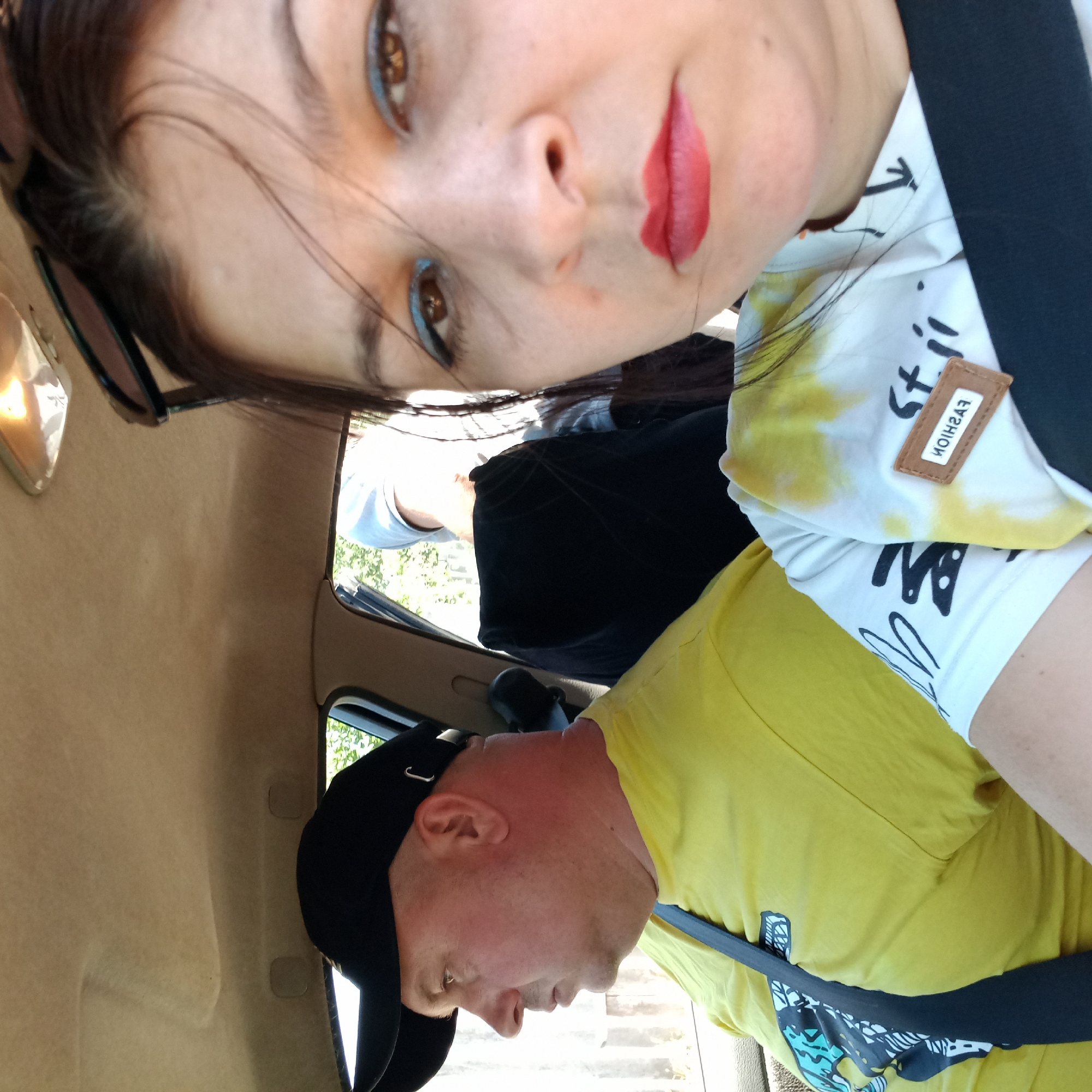 Two people are visible inside a car. In the foreground on the right, a woman with dark hair and red lipstick is looking at the camera. She is wearing a white t-shirt with yellow tie-dye elements and black graphics, and a black seatbelt. In the background on the left, a man with a black baseball cap and a yellow t-shirt is looking out the window. He is also wearing a black seatbelt. The interior of the car appears to be tan. Outside the window, greenery and a wooden fence are visible. There is no indication that this image was taken in Klyntsi, Ukraine.