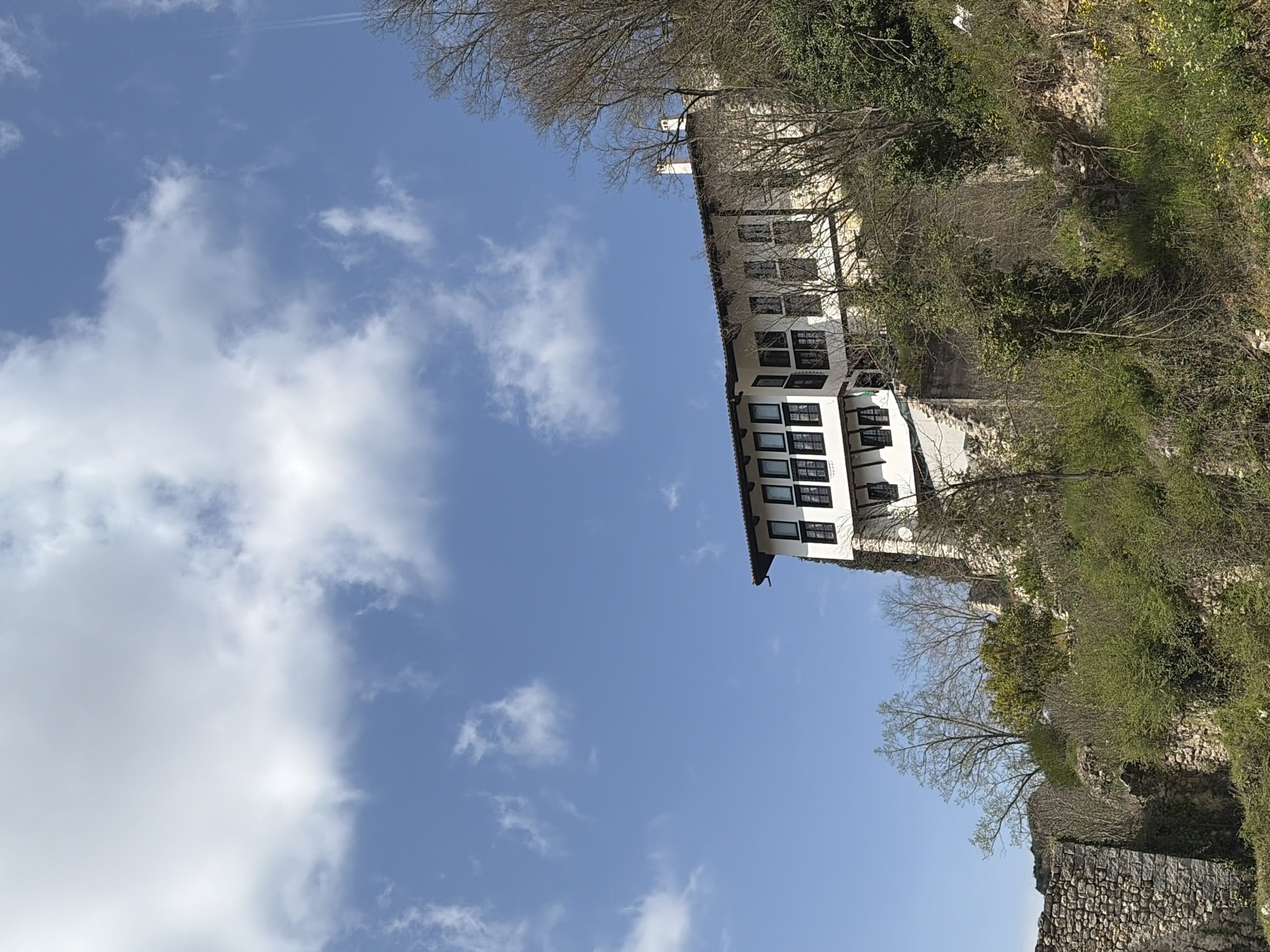 The image shows a multi-story building with white walls and dark-framed windows in an environment that includes a blue sky with scattered white clouds and vegetation including trees and greenery along a stone wall structure. The exact location in the city of Unknown, Unknown, cannot be determined from the image. There are no people visible in the image. The building appears to be situated on a slope or hill, with vegetation growing around it.