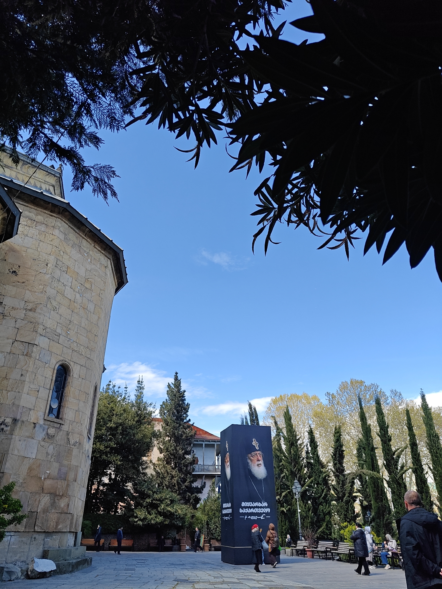 This daytime photograph captures a public square or courtyard in Tbilisi, Georgia, under a clear blue sky with a few wispy clouds. The scene is framed by dark, dense tree foliage from overhead on the top and left, contrasting with the bright sky.
On the left, a prominent stone building, likely a church or cathedral, features a rounded exterior with arched windows. Its facade is constructed from light-colored, rough-hewn stone blocks. In the mid-ground to the right, a tall, rectangular black pillar stands as a central object. This pillar displays large portraits of a man with a white beard and a religious headdress, identifiable as Ilia II, Catholicos-Patriarch of All Georgia. Below his image, text in the Georgian alphabet is clearly visible, including phrases like "უწმინდესი და უნეტარესი" (His Holiness and Beatitude) and "საქართველოს" (of Georgia).
The square is paved, and several people are present, either walking or sitting on scattered wooden benches. Their attire, including jackets and coats, suggests mild to cool weather. In the background, tall, slender evergreen trees (cypresses) are intermingled with deciduous trees displaying vibrant light green leaves, indicative of spring or early summer. A building with a red roof and balconies can be seen nestled among the trees.
The atmosphere appears calm and peaceful, with individuals enjoying the public space. The time of day seems to be late morning or early afternoon, with ample natural light. The distinct architecture, religious imagery, and Georgian script firmly place the setting in Tbilisi, Georgia.