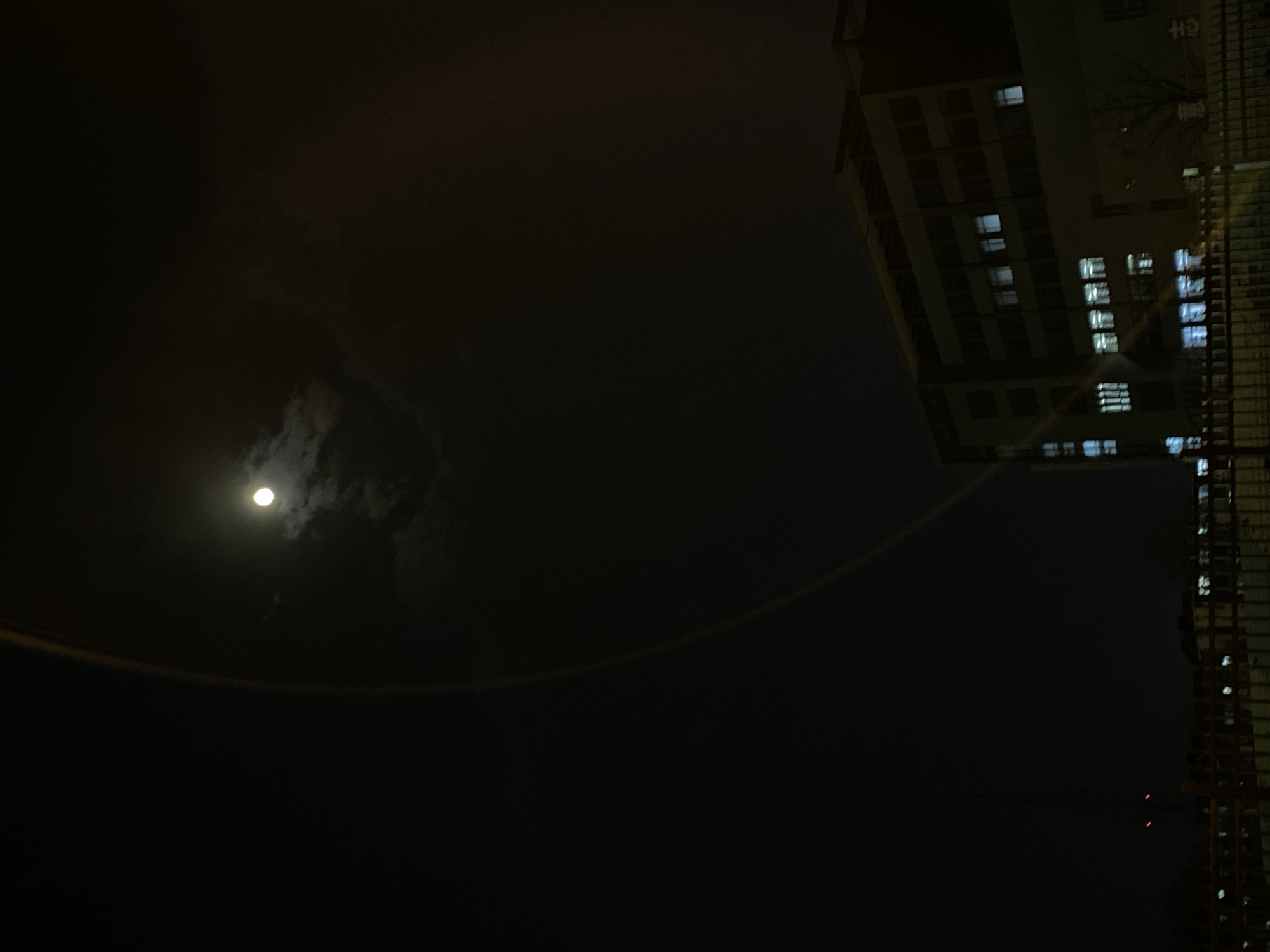 The image depicts a night scene in Zhongtun, China, showcasing a brightly lit moon partially obscured by clouds in the upper left quadrant. A large, multi-story building occupies the right side of the frame, its windows illuminated with artificial light. The sky is dark and the city atmosphere is obscured by shadow and darkness. A thin, curving line stretches across the lower portion of the sky.