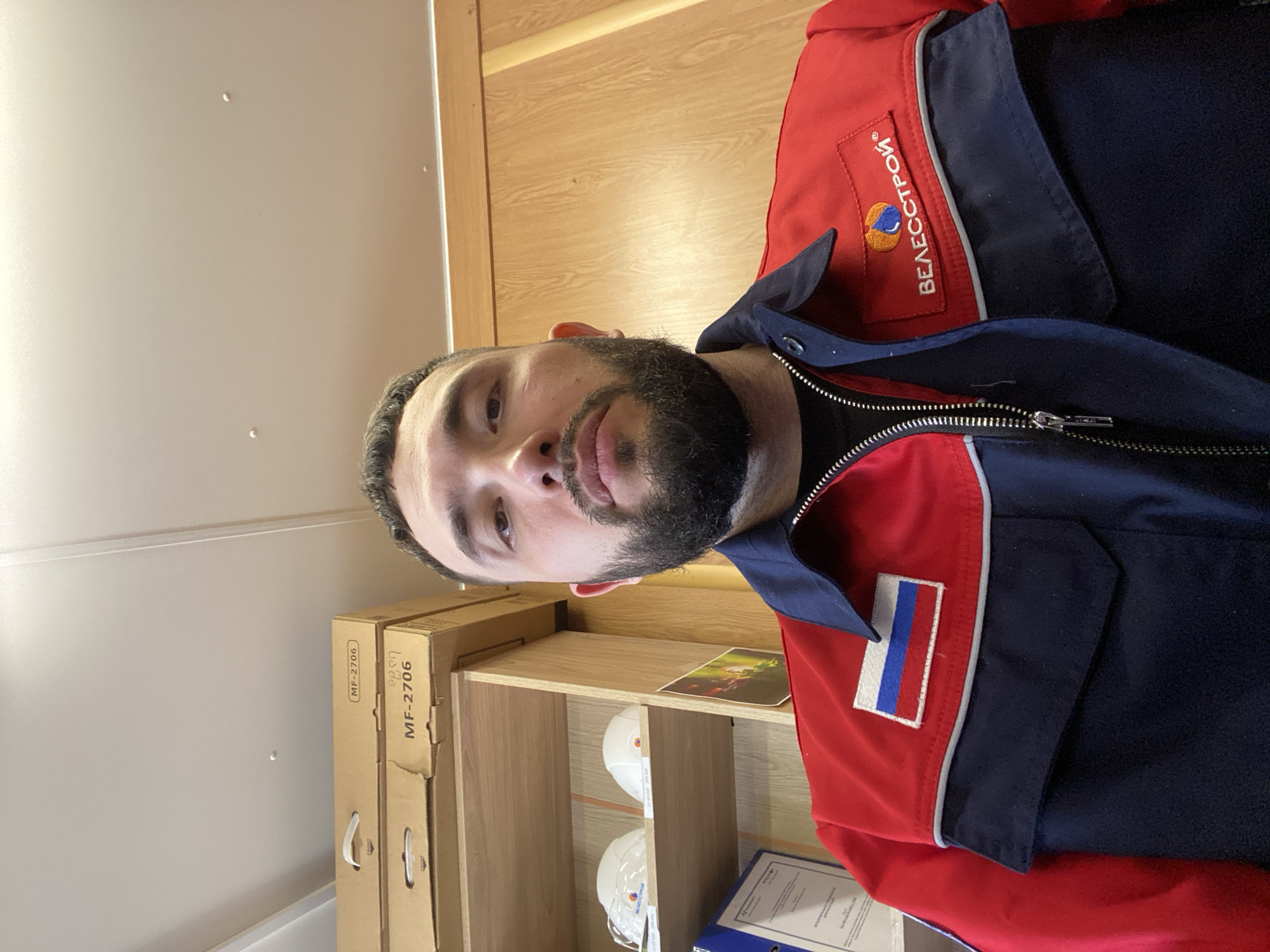 An image, rotated 90 degrees clockwise, shows a man indoors, likely in an office or workroom in Luzhitsy, Russia. The man, with dark hair and a beard, looks directly at the camera with a neutral expression. He is wearing a red and navy blue work jacket with a zipper down the front.
On the left chest of his jacket, there is an embroidered logo that reads "ВЕЛЕССТРОЙ" (VELESSTROY) in Cyrillic, accompanied by an abstract orange and blue graphic. On the right arm of the jacket, there is a patch resembling the Russian flag, with vertical stripes of white, blue, and red.
Behind the man, a light-colored wall and wooden shelving are visible. On the shelves, several items are stored: cardboard boxes, two white hard hats, and a blue binder. One of the cardboard boxes has the text "MF-2706" printed on it. A small, colorful picture or document is also placed on one of the shelves. The indoor lighting appears bright, suggesting it is daytime.