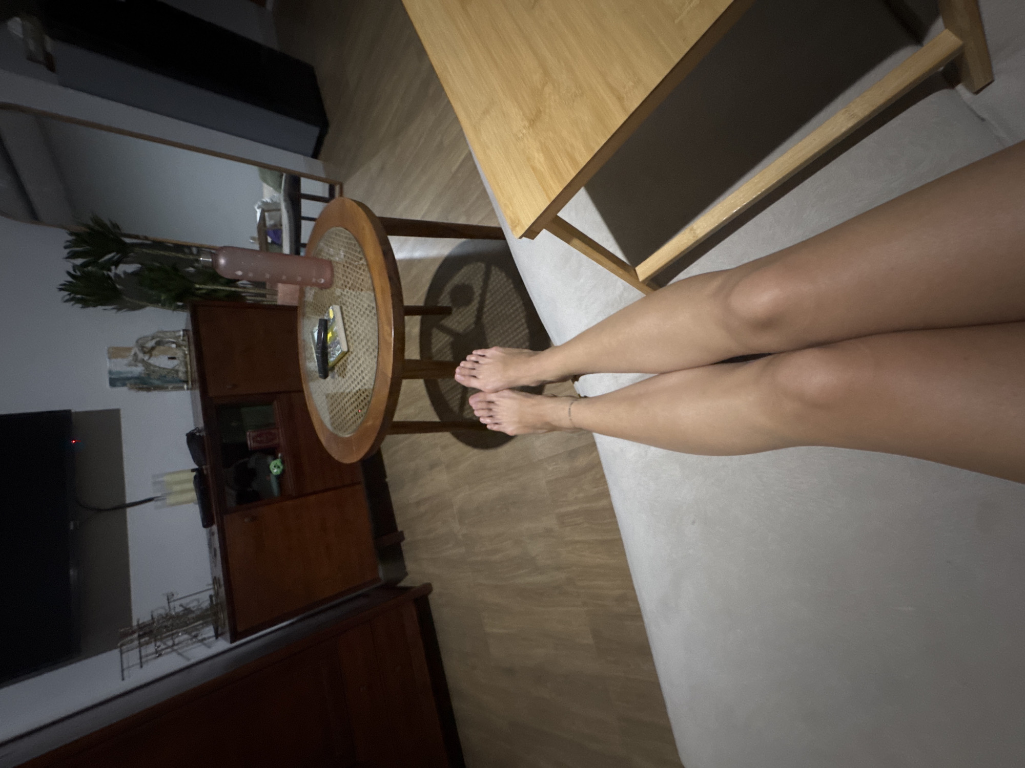 A low-angle, eye-level medium shot captures a person's legs and feet from the knees down, resting on a light gray, possibly upholstered surface, in what appears to be a living room in North Kuta, Indonesia. The scene is dimly lit, with warm ambient light casting shadows.
The person's legs are tanned and smooth, with a thin, delicate anklet visible on the left ankle. The feet are bare, with toes pointing upwards and slightly outward. The right foot is partially obscured by the left.
In the background, a round, dark wood coffee table with a woven top is positioned to the left of the person's legs. On the table, a book, a remote control, and a pink water bottle are visible. To the left of the coffee table, a dark wood cabinet with glass-fronted doors stands against a light-colored wall. On top of the cabinet, an array of metal racks holding various items, a television mounted on the wall, and a few decorative objects are present.
Further into the background, a tall, dark-framed mirror leans against the wall, reflecting a portion of the room, including a potted plant. To the right, a light-colored wooden tray table is angled towards the foreground, casting a dark shadow. The flooring is a warm-toned wood laminate. The overall atmosphere suggests a relaxed, domestic setting.