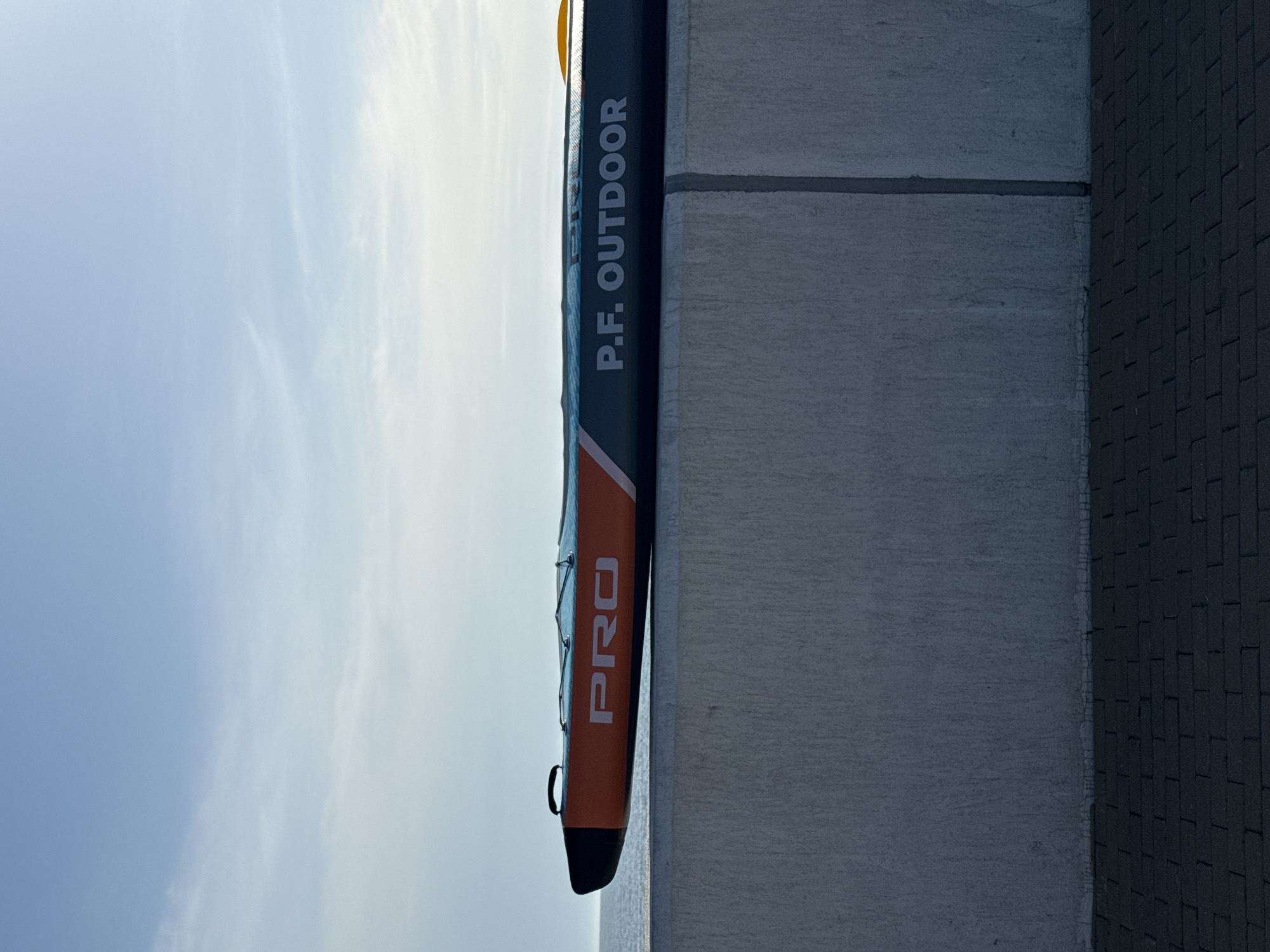 A vertical shot presents a product leaning against a concrete structure, likely a wall or pillar, with a glimpse of the sky in the background. The product appears to be a paddleboard or inflatable kayak, branded with "P.F. OUTDOOR" printed vertically in white letters on a dark background. The bottom part of the product has "PRO" written in large, white letters on an orange surface. The concrete structure exhibits a smooth, gray texture. The background shows a pale blue sky with light clouds. The black brick wall to the right is located at the intersection with Olympic Avenue and Vzletnaya street in Sirius, Russia.