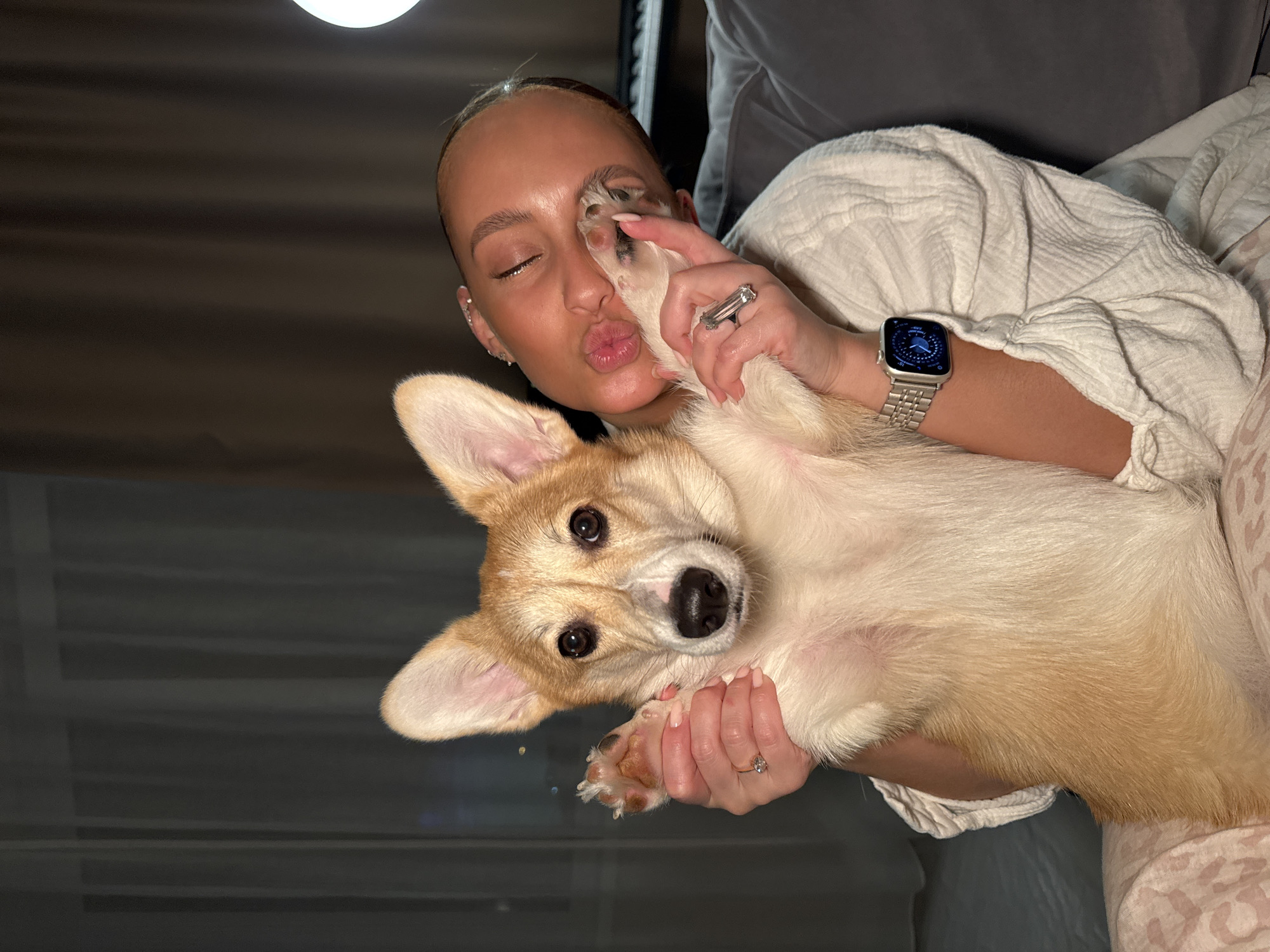 A young woman in a white robe is lying down and holding a corgi dog. She is pouting and holding the dog's paw to her face. The dog is looking at the camera. The location is unknown; there is no indication it is in the city of Chaikovsky, Russia.