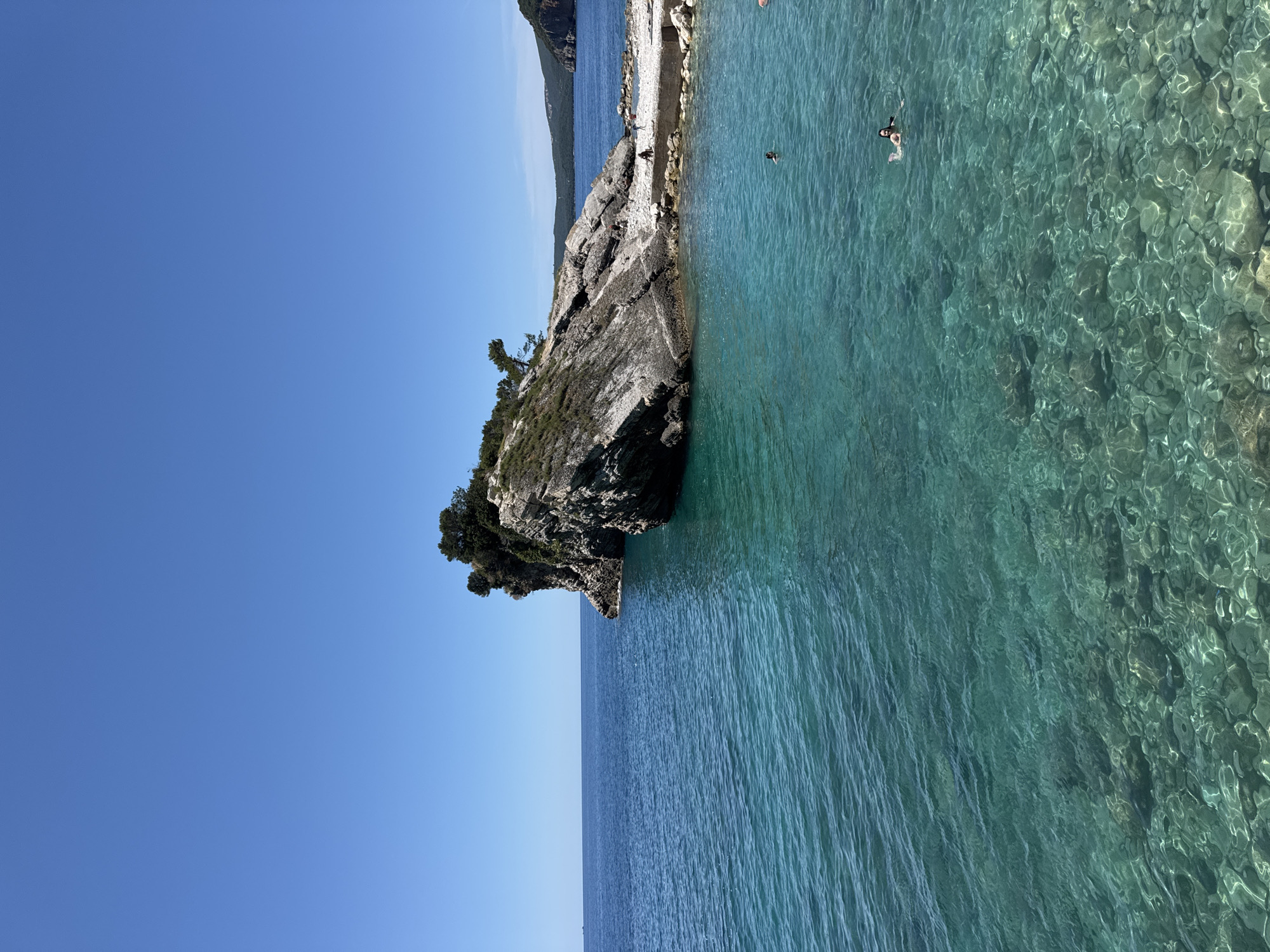 The image depicts a serene coastal landscape in Budva, Montenegro, bathed in bright sunlight under a clear, expansive blue sky. The scene is dominated by the calm, crystal-clear turquoise waters of the Adriatic Sea, which reveal the rocky seabed beneath its surface, especially closer to the shore. Ripples on the water reflect the light, creating shimmering patterns.
On the left side of the frame, a rugged, dark grey rocky outcrop or small headland extends into the sea. This natural formation is sparsely adorned with small green trees and bushes, adding touches of verdant life to the stone. Beyond this prominent rock, in the mid-ground, a larger landmass, likely a hill or mountain, covered in dense green vegetation, stretches along the distant coastline. A narrow, light-colored strip, possibly a small beach or rocky path, can be seen at the base of this larger landmass.
The activity in the image focuses on leisure. One person is visible swimming or snorkeling in the pristine, shallow waters near the rocky shore, enjoying the transparent sea. A couple of other small figures appear to be standing or walking on the distant shoreline.
The time of day appears to be midday or early afternoon, characterized by the intense sunlight and the absence of clouds. The weather is perfectly clear and sunny, contributing to the vibrant hues of the water and sky. The overall atmosphere is one of peaceful natural beauty and ideal conditions for seaside enjoyment. There is no visible text in the media file.