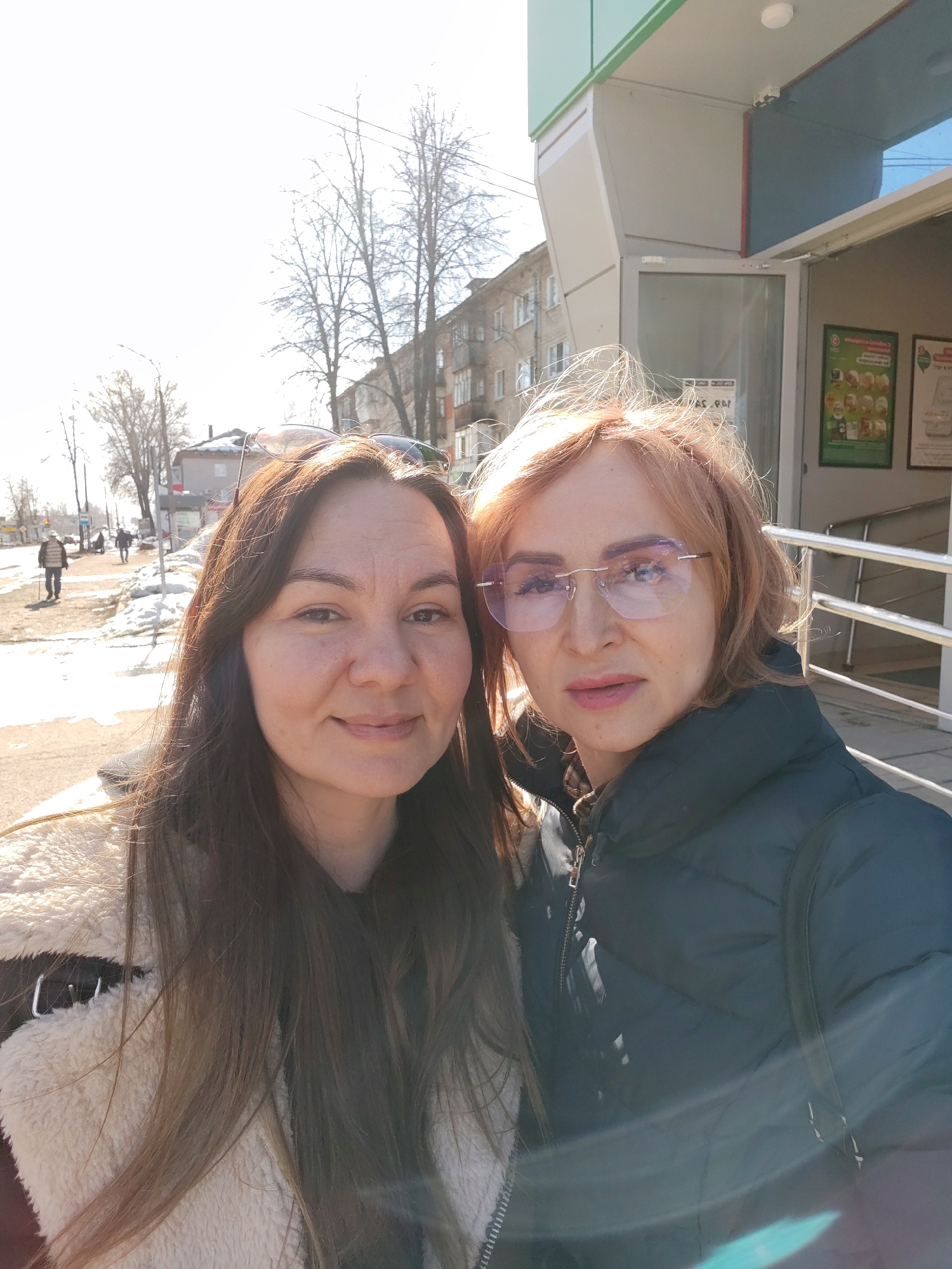 This image depicts two adult females positioned in the foreground, facing the camera for what appears to be a selfie on a sunny day. The scene is located on a city street in Chaikovsky, Russia, specifically in front of the "ПарСыр" (ParSyr) cheese shop at Ulitsa Lenina, 50B.
The female on the left has long, dark brown hair, which is parted down the middle. She wears a light-colored, likely cream or off-white, fuzzy jacket or vest over a dark garment, and a pair of sunglasses is perched on top of her head. Her facial expression shows a slight smile. The female on the right has shorter, light reddish-brown hair and wears rimless eyeglasses with a subtle blue tint. She is dressed in a dark, possibly navy or black, puffer jacket. Her expression is neutral.
In the immediate background to the right, a modern commercial building features a facade with alternating green and light grey panels. A glass entrance door is partially open, revealing promotional posters with green branding on an interior wall. A silver-colored handrail extends from the building's exterior. A white circular sensor or light fixture is mounted above the entrance.
The distant background on the left reveals a street with visible patches of snow on the ground and along the sidewalk, indicating cold weather. Bare deciduous trees line the street, and multi-story residential buildings are visible further in the distance. Several pedestrians are present on the sidewalk, including one on the far left who appears to be using a walking stick. The sky is clear, and strong sunlight is evident, creating lens flare in the image.