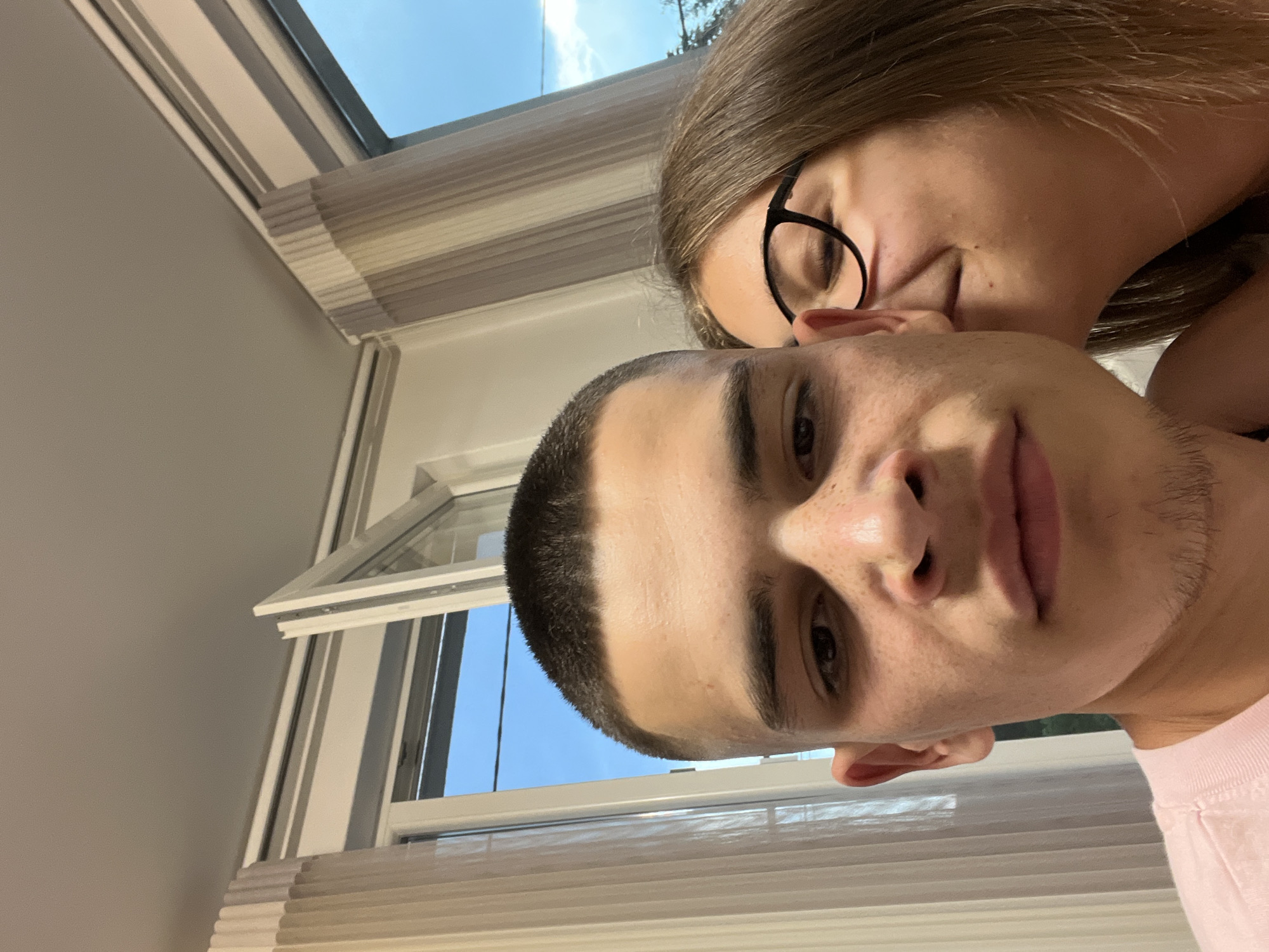An intimate close-up photograph depicts a male and a female in Karavelovo, Bulgaria. In the foreground, a young male with a short buzz-cut hairstyle, prominent freckles across his nose and cheeks, and faint stubble on his chin, gazes directly into the camera. He is wearing a light pink crew-neck t-shirt. Partially obscured behind him and to his right, a female with long light-brown hair, wearing black-framed glasses, leans in with her face close to the male's right ear, appearing to whisper or kiss. Her right eye is closed, and a subtle smile is present. The background reveals an indoor setting with a large window structure. Light-colored vertical blinds or curtains partially cover the window on the left and right. A central section of the window is open, providing a view of a bright blue sky with some white clouds and a thin, dark horizontal line (possibly a wire) in the distance. Dark green foliage is visible through the lower right portion of the open window. The ceiling above is a plain, light gray surface.