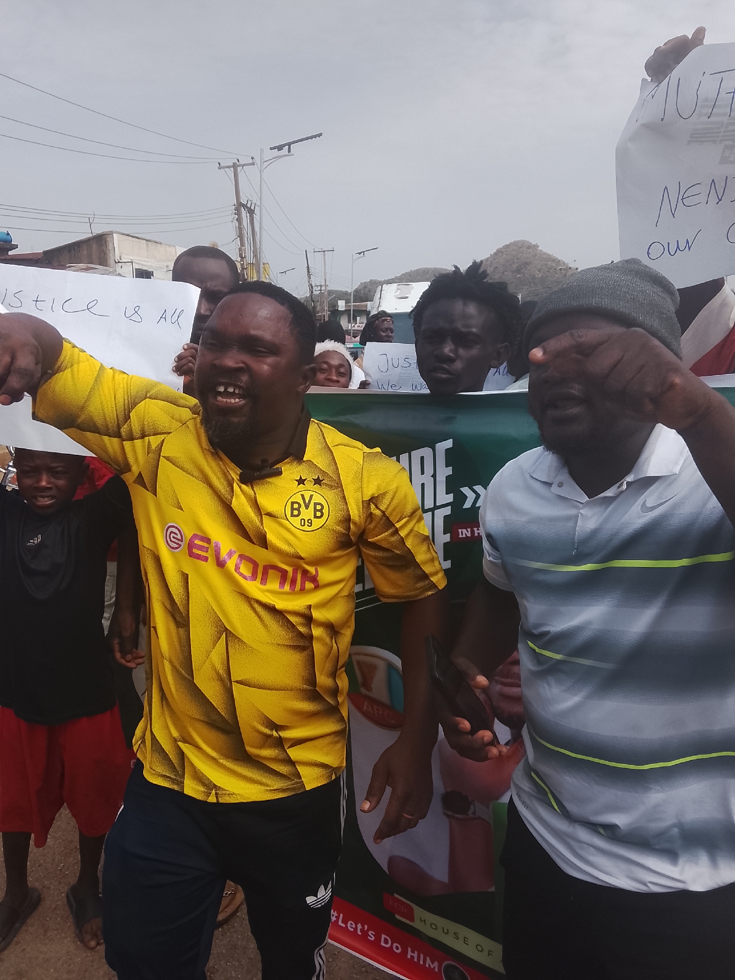 The image shows a group of people, likely in Gwong, Nigeria, participating in a protest or demonstration.
The main subject is a man in a yellow Borussia Dortmund soccer jersey. He is gesturing and appears to be shouting. Next to him is another man, partially covering his face with his hand and wearing a gray striped shirt and a gray beanie. Other people are visible behind them, some holding signs with writing on them, including "Justice is All," "MUTH," "NEN," and "Our C." A banner is partially visible with the words "RE", "E", "IN H", "APC", "FOR," "HOUSE OF", and "#Let's Do HIM."
The environment shows a street scene with buildings, utility poles, and some greenery in the background. The weather appears overcast.