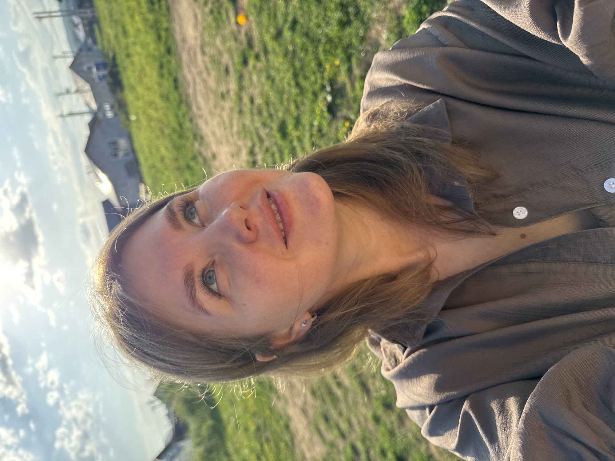 A photograph shows a person with fair skin and light brown hair wearing a collared shirt. The shirt is brown with white buttons. The person's gaze is directed upwards. The background is out of focus, but it appears to be a grassy area with some buildings visible in the distance under a partially cloudy sky. The location is Ферма, Russia.