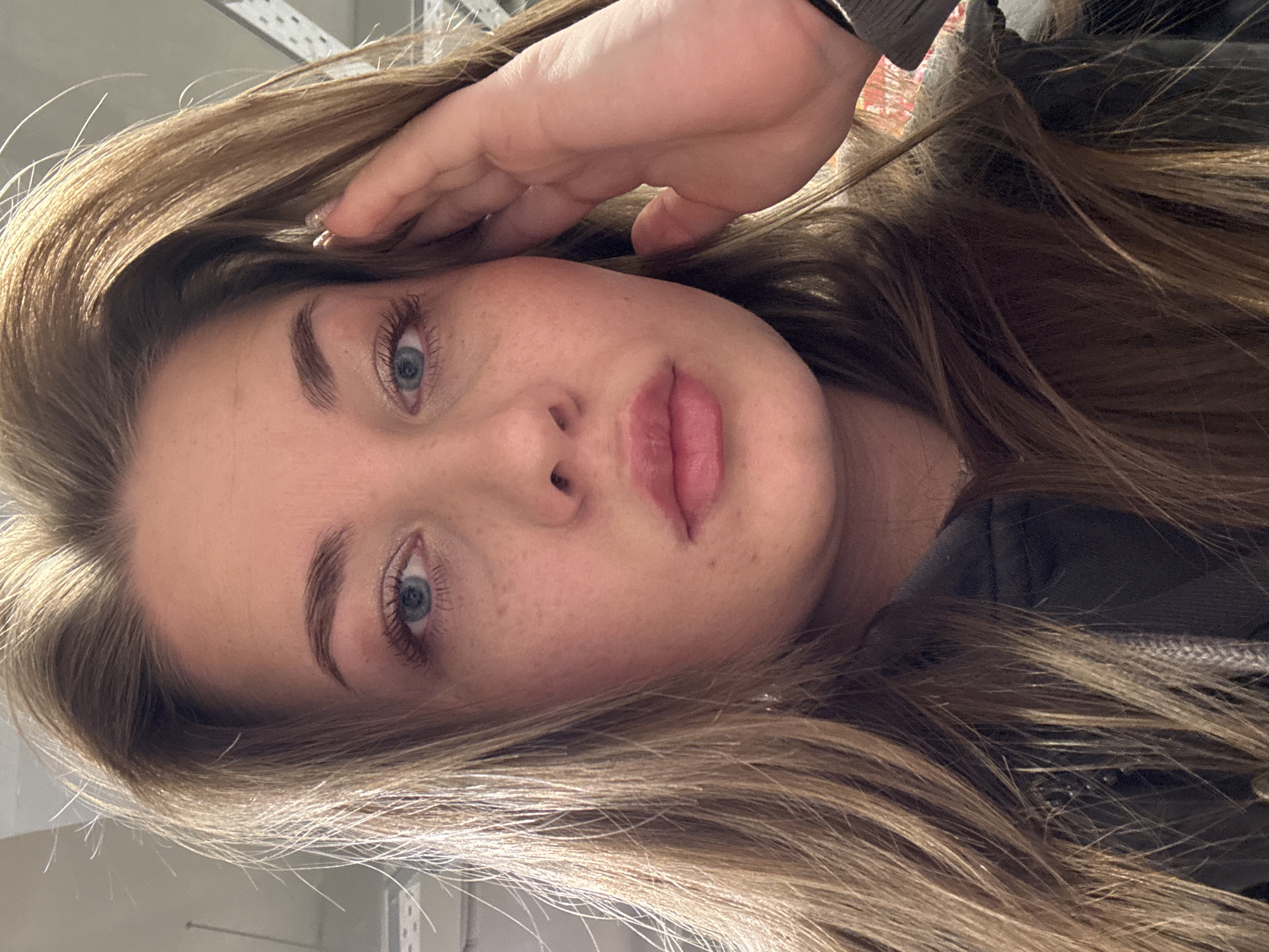 A close-up portrait of a young woman with light brown hair, blue eyes, and freckles on her face. She is looking directly at the camera with her right hand slightly raised towards her forehead. She has pinkish lips. Her hair is long and loose, falling around her face and shoulders. She is wearing a dark-colored jacket or shirt. The background is slightly blurred and appears to be an interior space, with a hint of a metal structure visible in the upper left corner. Location cannot be determined as Chaikovsky, Russia.