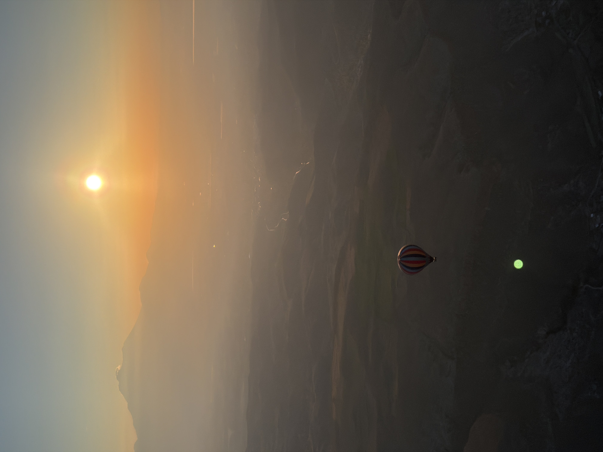The image captures an aerial view of a vast, mountainous landscape at either sunrise or sunset, creating a tranquil and atmospheric scene. The sun, a brilliant white-yellow orb, is positioned on the left side of the frame, casting a warm, golden glow and streaks of light across the hazy sky. The sky transitions from a pale blue-grey on the far left to soft oranges and browns, suggesting the scattering of light through atmospheric haze.
The terrain below consists of dark, rugged valleys and hills, illuminated faintly by the distant light, revealing subtle contours and textures across the expansive landscape. The overall mood is one of serenity and awe.
A single hot air balloon, distinctively patterned with horizontal stripes of red, blue, and white, floats gracefully in the middle-right portion of the image. Its relatively small size against the immense backdrop underscores the vastness of the environment. To the right of the balloon, a bright, circular green light or orb is visible, which appears to be a lens flare or a distant light source, contrasting with the warm natural light.
Given the context of Yeşilhisar, Turkey, this scene likely depicts a hot air balloon flight over the Cappadocia region, renowned for its unique geological formations and sunrise balloon experiences. No text is visible in the image.