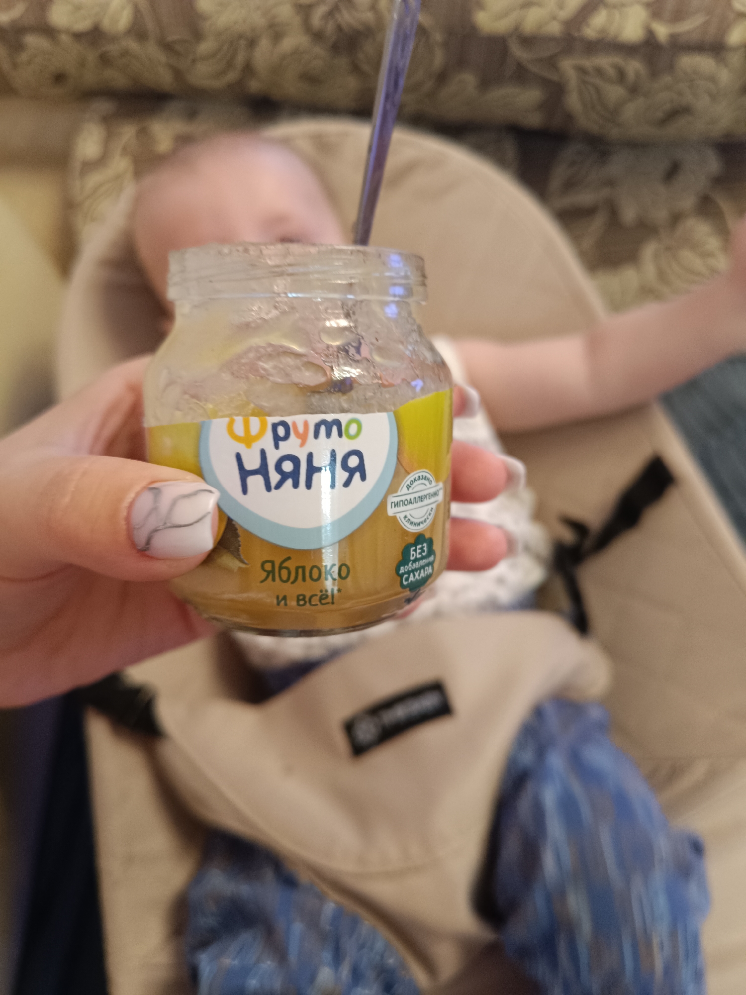 A hand holds a jar of "Fruto Nyanya" apple baby food, with the text "яблоко и всё!" (apple and that's it!) on the label. A baby is lying in a baby carrier in the background. The text on the jar includes "ГИПОАЛЛЕРГЕННО" (hypoallergenic) and "БЕЗ добавления САХАРА" (no added sugar). No location in Tobolsk, Russia is evident.