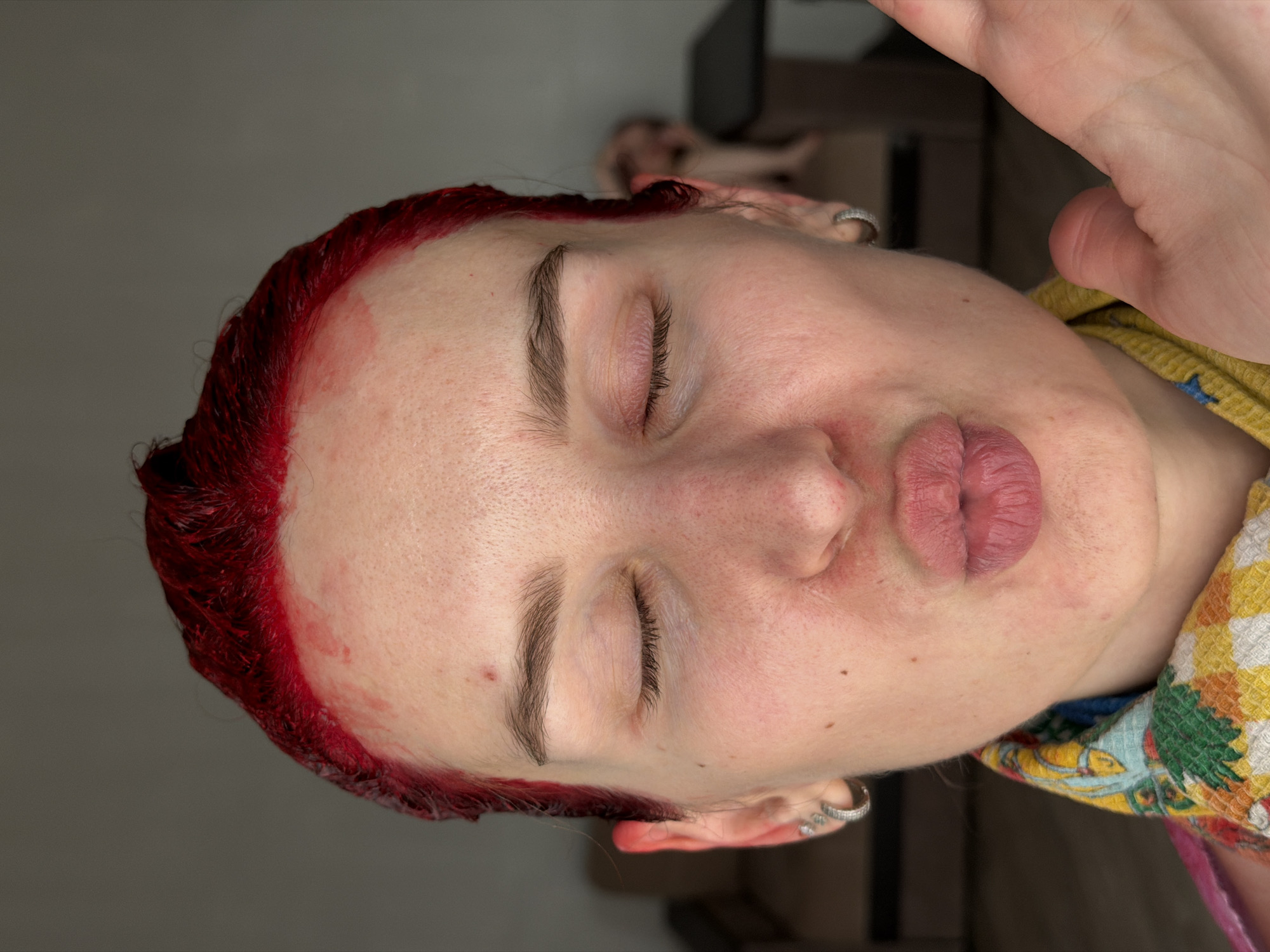 A close-up, indoor photograph presents a person with closed eyes and red hair, likely recently dyed as evidenced by the dye staining the forehead. The individual is positioned on their side, suggesting they are lying down. The background is blurred. A hand is visible on the upper right portion of the image. The location is not identifiable from this image.