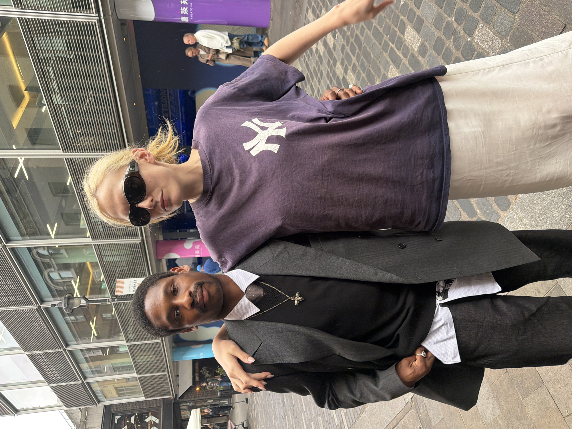 Two individuals are pictured in an outdoor urban setting. The background includes a building with architectural details and signage in English and other languages. Cobblestone pavement is visible.
A woman with blonde hair and sunglasses wears a purple t-shirt with a white New York Yankees logo and a light-colored skirt. She has rings on her fingers and her arm is outstretched.
A man in a suit, wearing a black shirt with a white collar and a cross pendant, is partially covered by the woman and has her arm draped around his shoulder. He is wearing a ring.
The location appears to be in the city of London, United Kingdom.