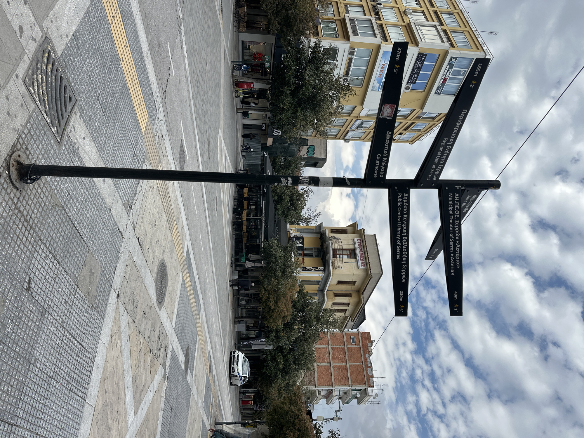 A directional signpost stands on a tiled sidewalk in Serres, Greece. The sign indicates the locations of several key landmarks. The courthouse is one, also the Public Central Library of Serres, the Regional Unit of Serres, the Municipal Theater of Serres, and the Town Hall. The signs include distances, in meters and the estimated walking time. The background shows buildings with commercial signage and trees. A parked police vehicle is visible in the street. The sky is overcast.