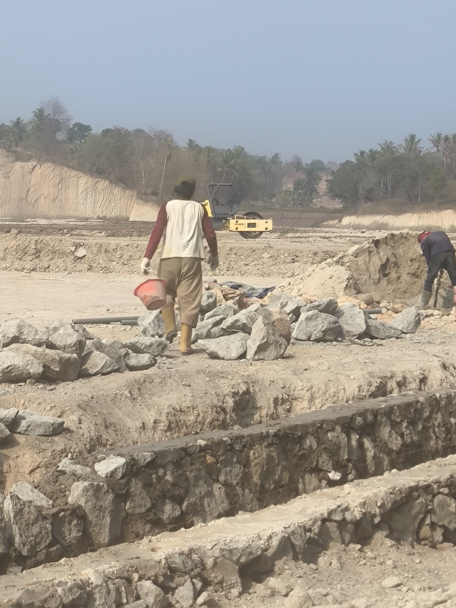 In the city of Cilegon, Indonesia, a construction site is depicted under a clear sky. The scene is dominated by raw earth and piles of rocks, suggesting an ongoing earthmoving or foundation-laying project. In the foreground, a worker, seen from behind, walks away from the camera, carrying a red bucket in their right hand. They are wearing a light-colored vest over a reddish long-sleeved shirt, loose-fitting beige pants, and yellow boots. To the right, another construction worker, wearing a dark jacket, dark pants, and a red hard hat, is bent over, seemingly working with a tool. In the mid-ground, a yellow steamroller is parked on the dirt. The background reveals tiered earthen banks and a line of tropical trees, including palm trees, indicating a warm climate. The overall impression is of an active construction zone in a potentially rural or developing area of Cilegon.