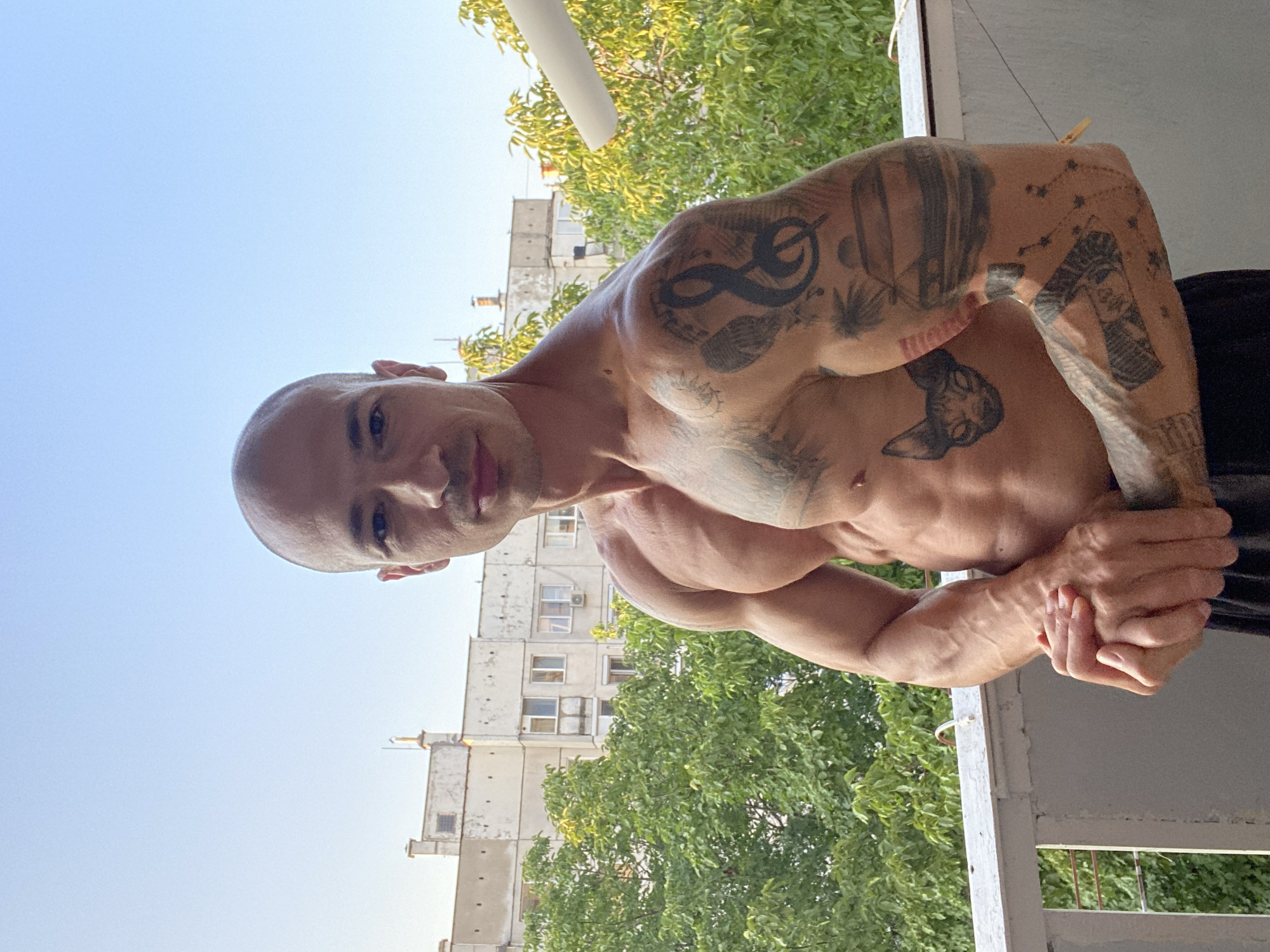 A muscular, bald man with tattoos on his arms and torso stands on a balcony in Pleven, Bulgaria. He is shirtless, wearing black shorts. Behind him, a multi-story apartment building with a weathered facade and green trees are visible. The sky is clear blue.